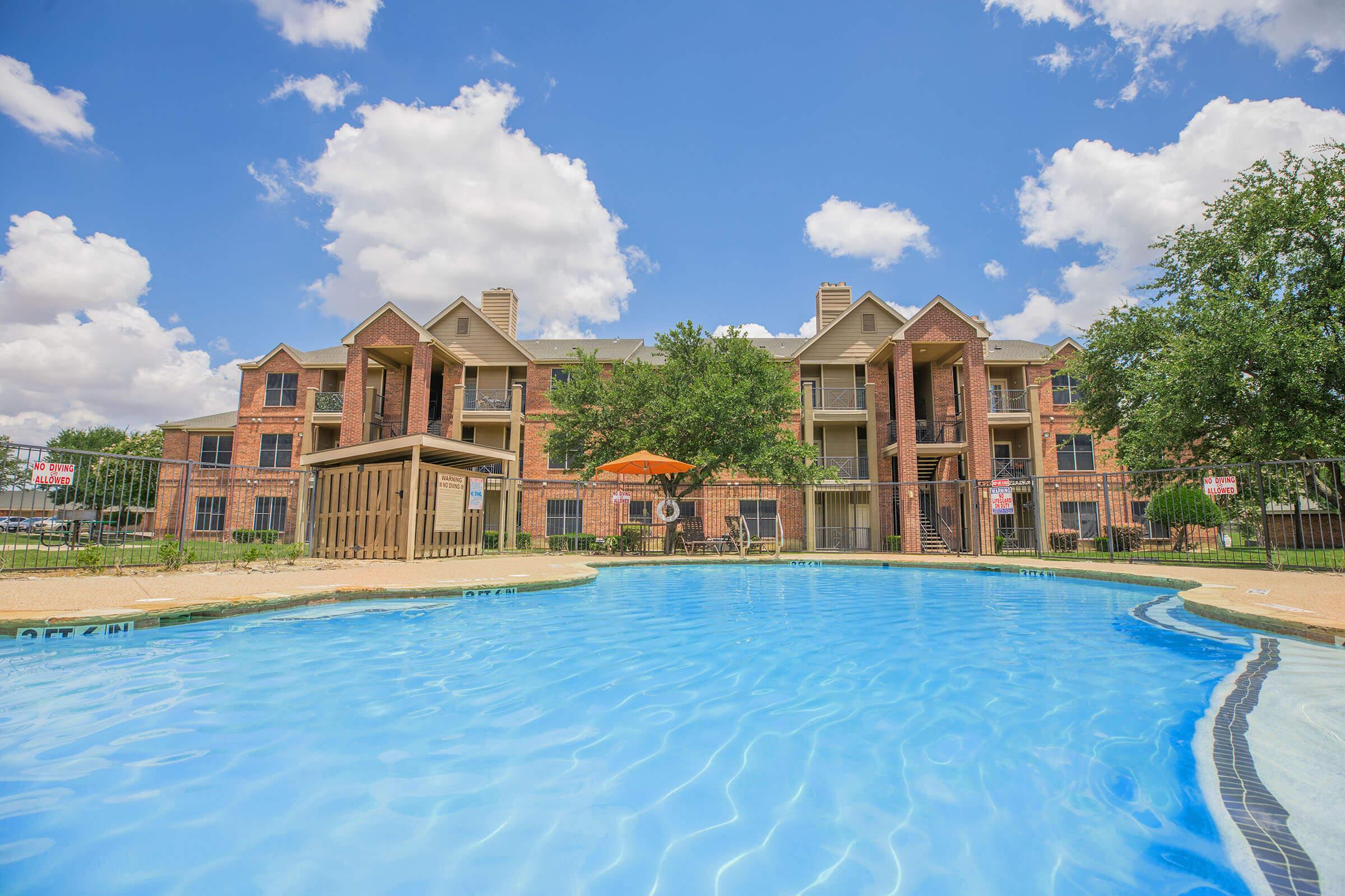 A bright, inviting swimming pool with clear blue water in the foreground, surrounded by a well-maintained outdoor area. In the background, a multi-story brick apartment building with balconies and lush green trees. A colorful umbrella provides shade near the pool. Fluffy clouds dot the blue sky.