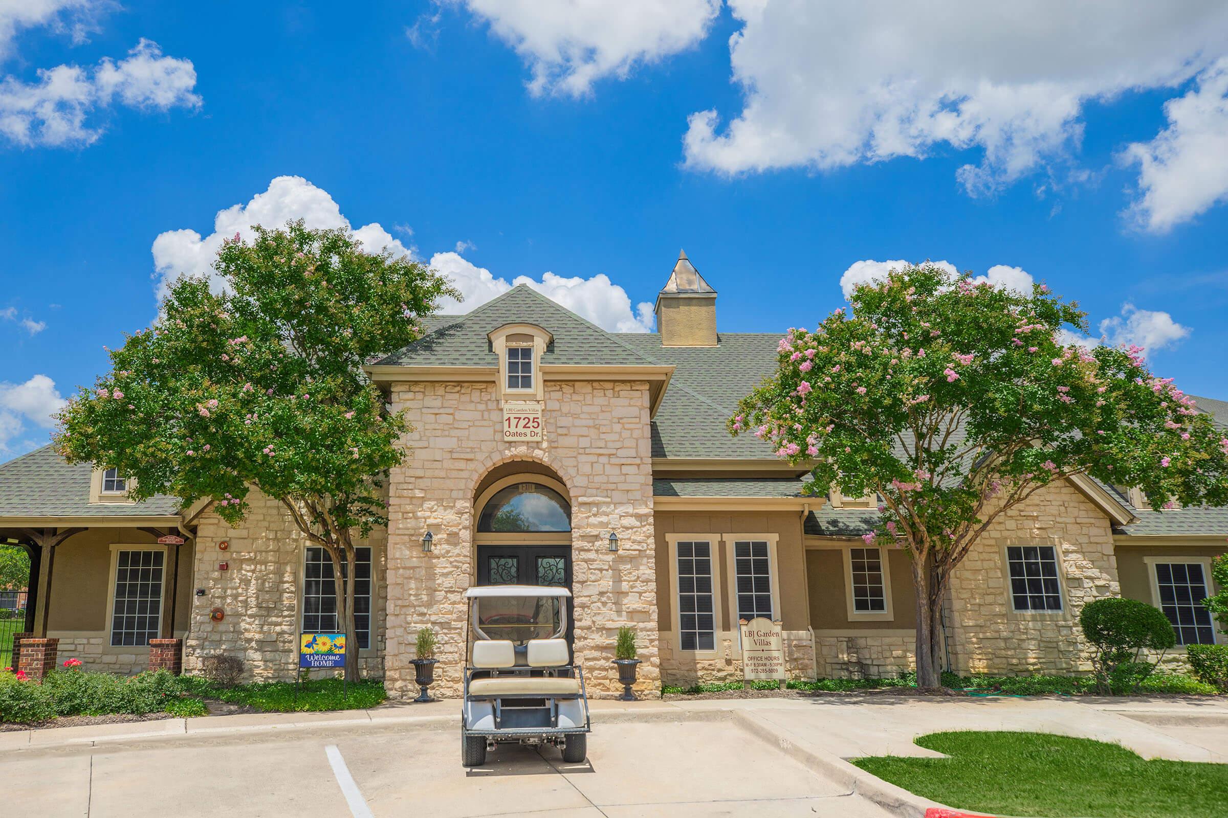 A well-maintained building featuring stone and stucco architecture, surrounded by lush green trees and shrubs. In the foreground, there is a parked golf cart. The sky is bright blue with fluffy white clouds, adding to the overall inviting atmosphere of the location.