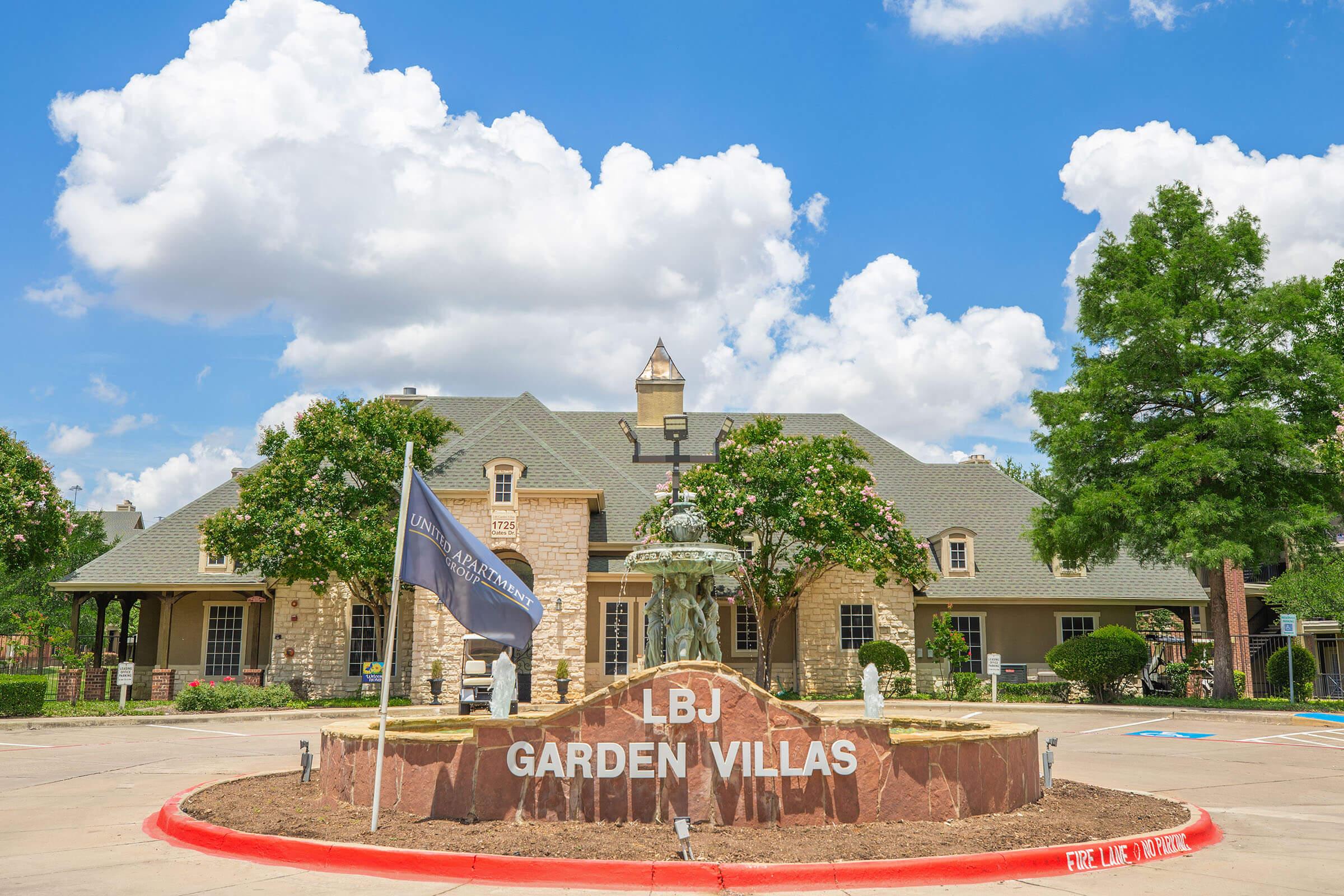 A welcoming entrance featuring a sign for LBJ Garden Villas surrounded by landscaped greenery. The building has a charming design with a peaked roof and stone accents, framed by large trees and bright blue skies with fluffy white clouds. A flag is also present in the foreground.