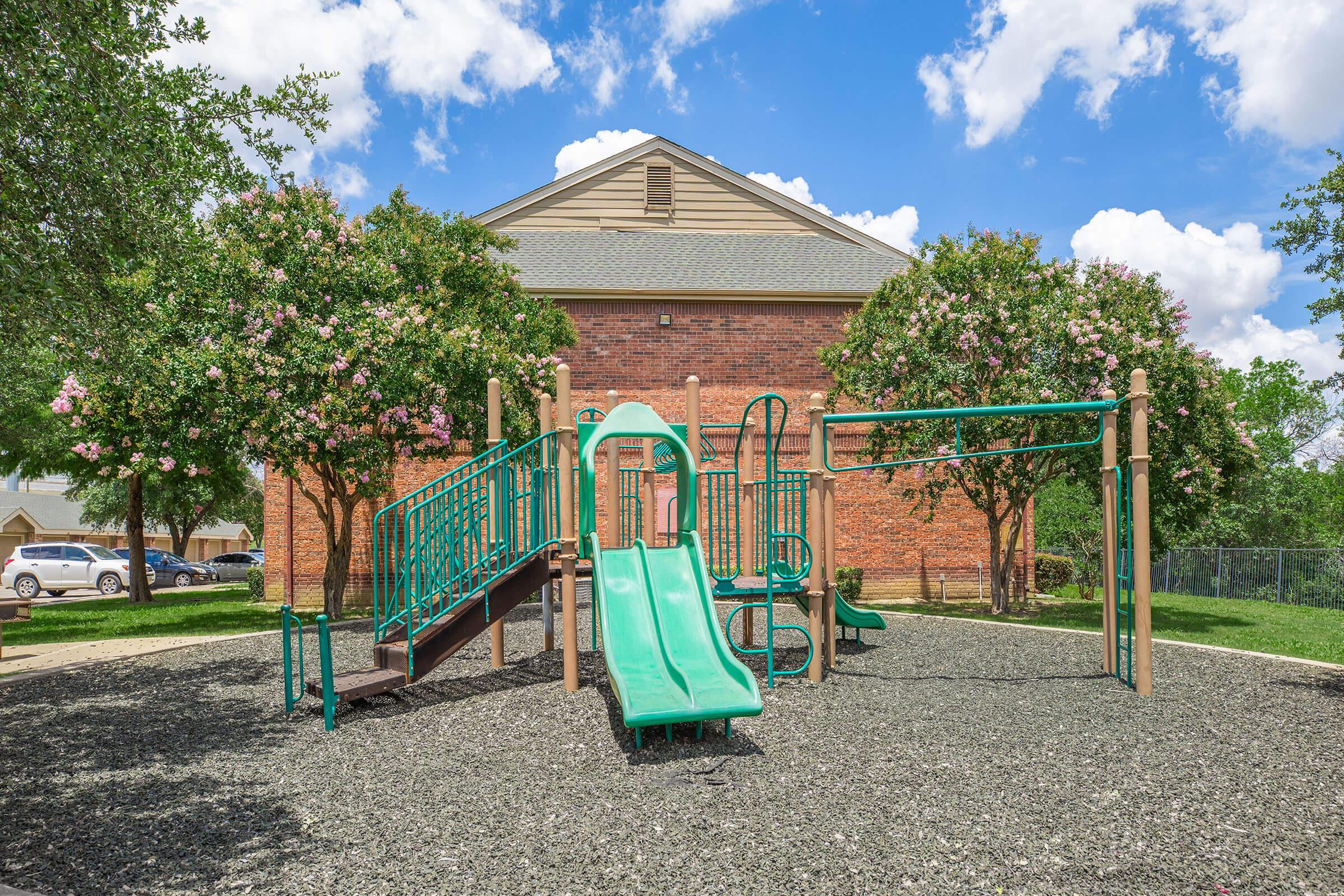 Playground scene featuring green slides and climbing structures set on a rubberized surface, surrounded by landscaped trees with pink flowers. A red brick building stands in the background under a bright blue sky with fluffy white clouds.