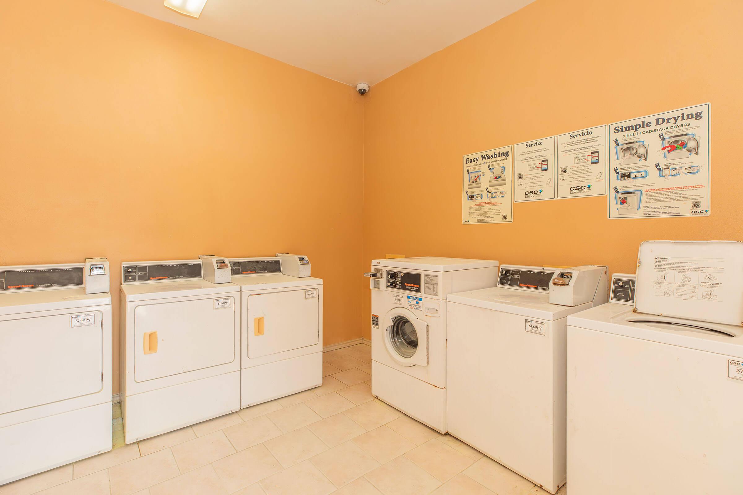 A laundry room featuring several white washing machines and dryers against a light orange wall. Instructional posters about washing and drying are displayed. The space is well-lit and tidy, with a tiled floor.