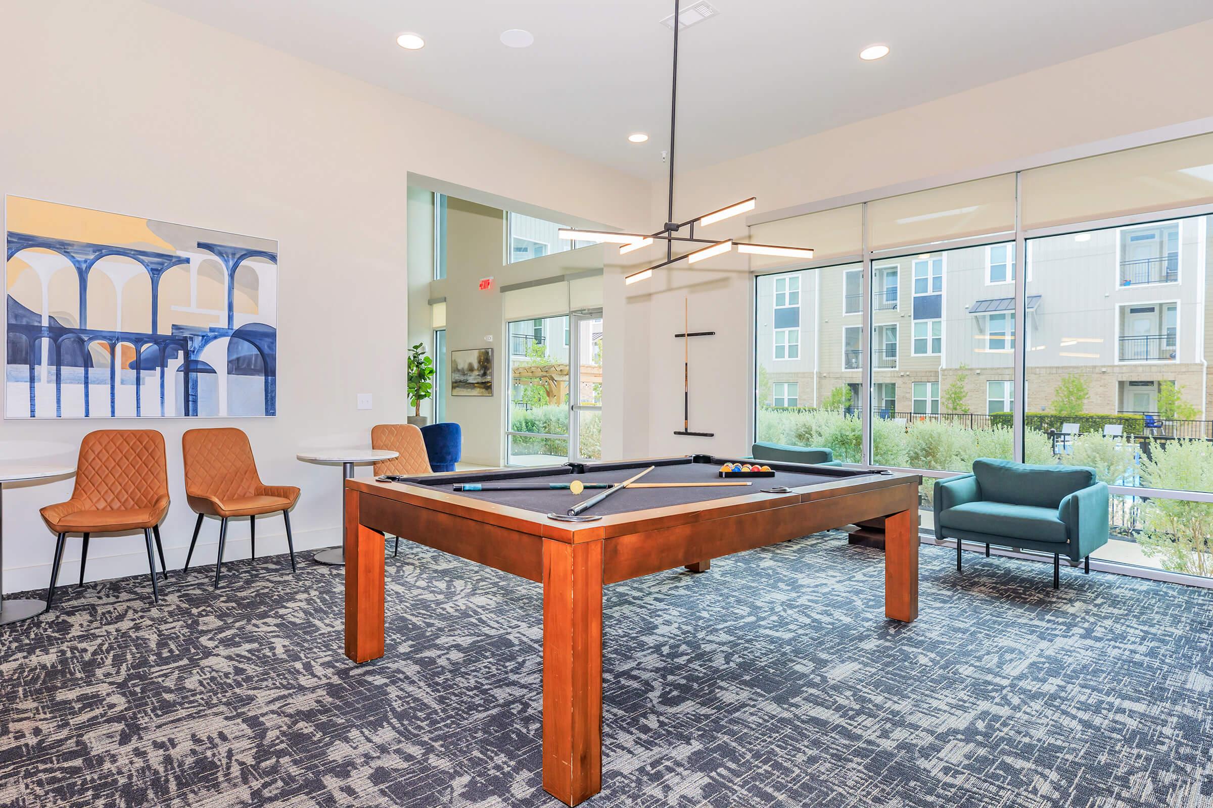 A modern recreational room featuring a pool table with colorful balls, surrounded by stylish seating. Large windows provide abundant natural light, and contemporary light fixtures hang above. The space has a sleek, inviting design with neutral colors and textured carpet.