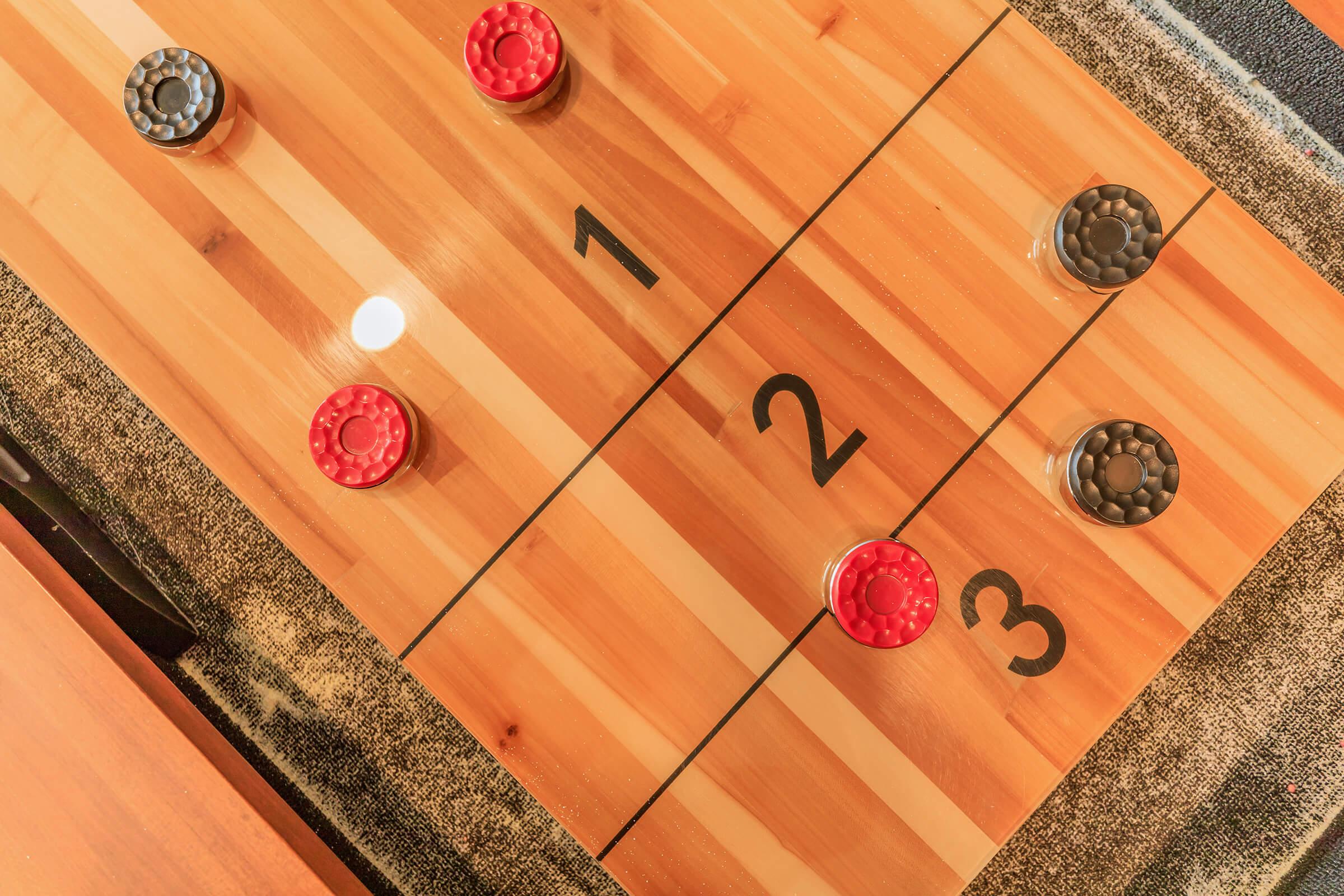 A close-up view of a wooden shuffleboard table with three numbered sections. There are two black discs and two red discs positioned on the board, with a smooth surface reflecting light. The background includes a dark cloth, enhancing the board's appearance.