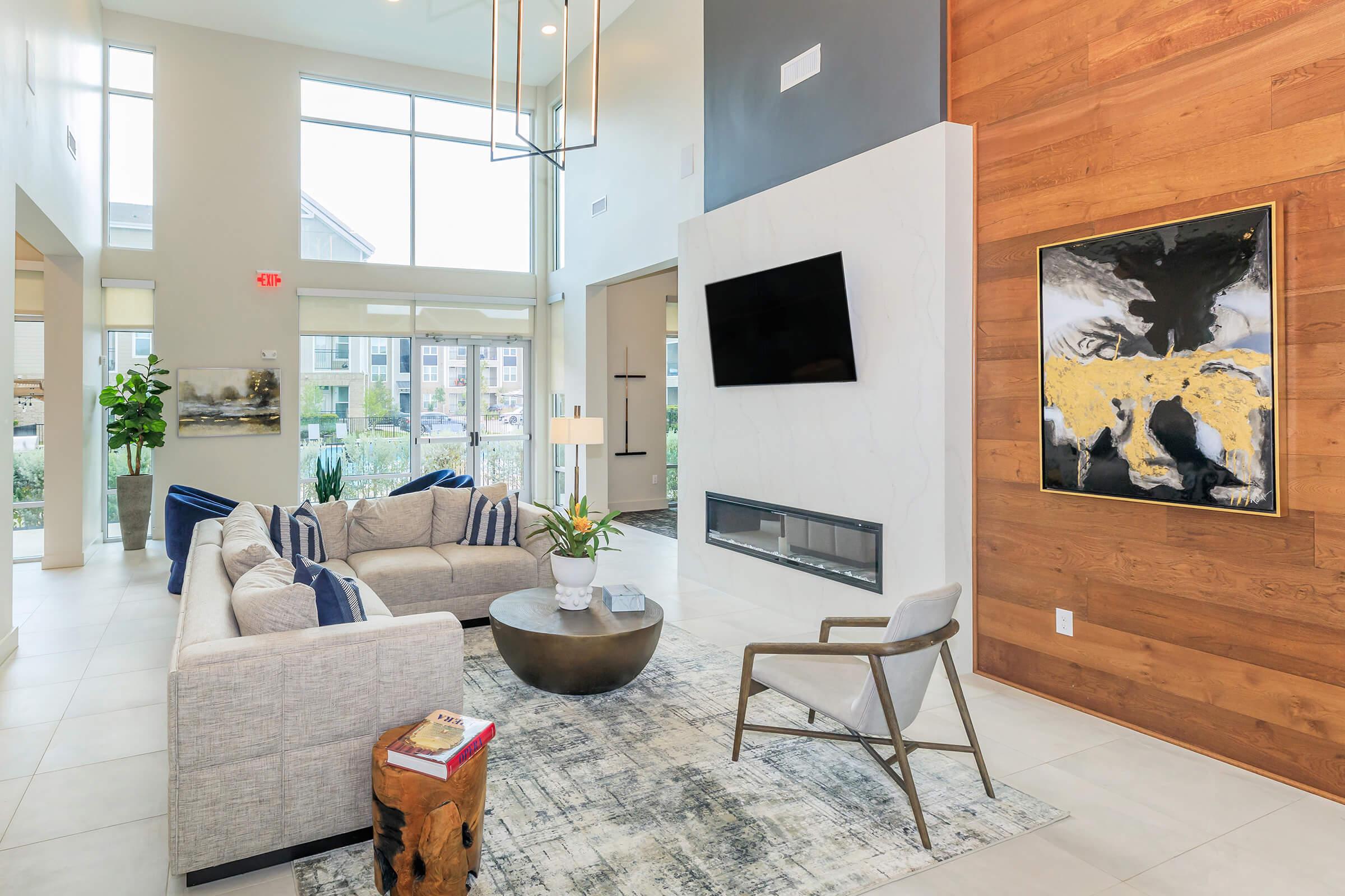 A modern living room featuring a light gray sectional sofa with blue accent pillows, a round coffee table, and a stylish armchair. The space has large windows allowing natural light, wooden accent walls, a mounted television, and contemporary artwork. A decorative plant adds a touch of greenery.