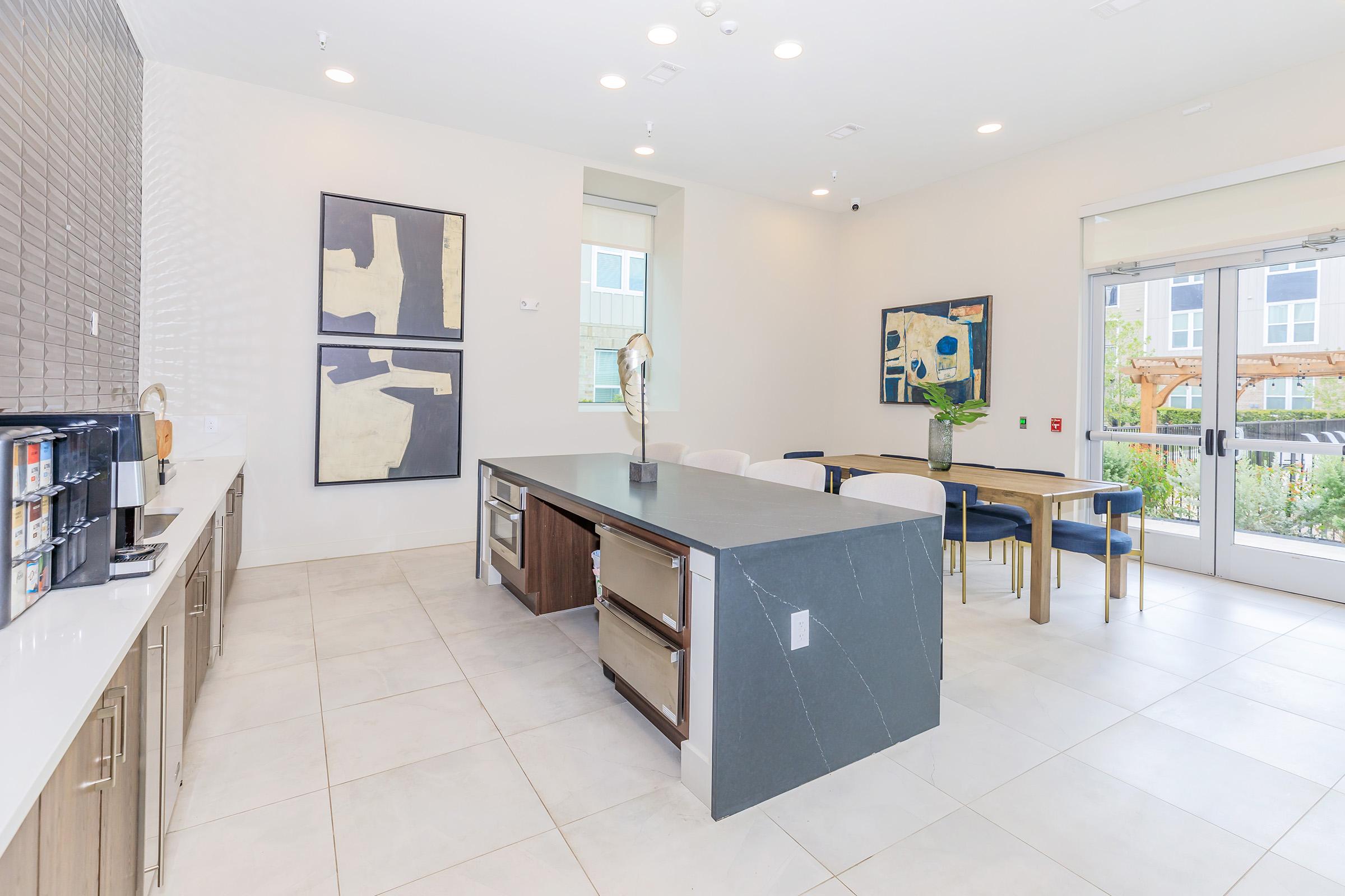 A modern communal space featuring a large island with a dark countertop, beige chairs, and a dining table. The walls are adorned with contemporary artwork, and there is a coffee station on one side. Large windows allow natural light to fill the area, enhancing the inviting atmosphere.