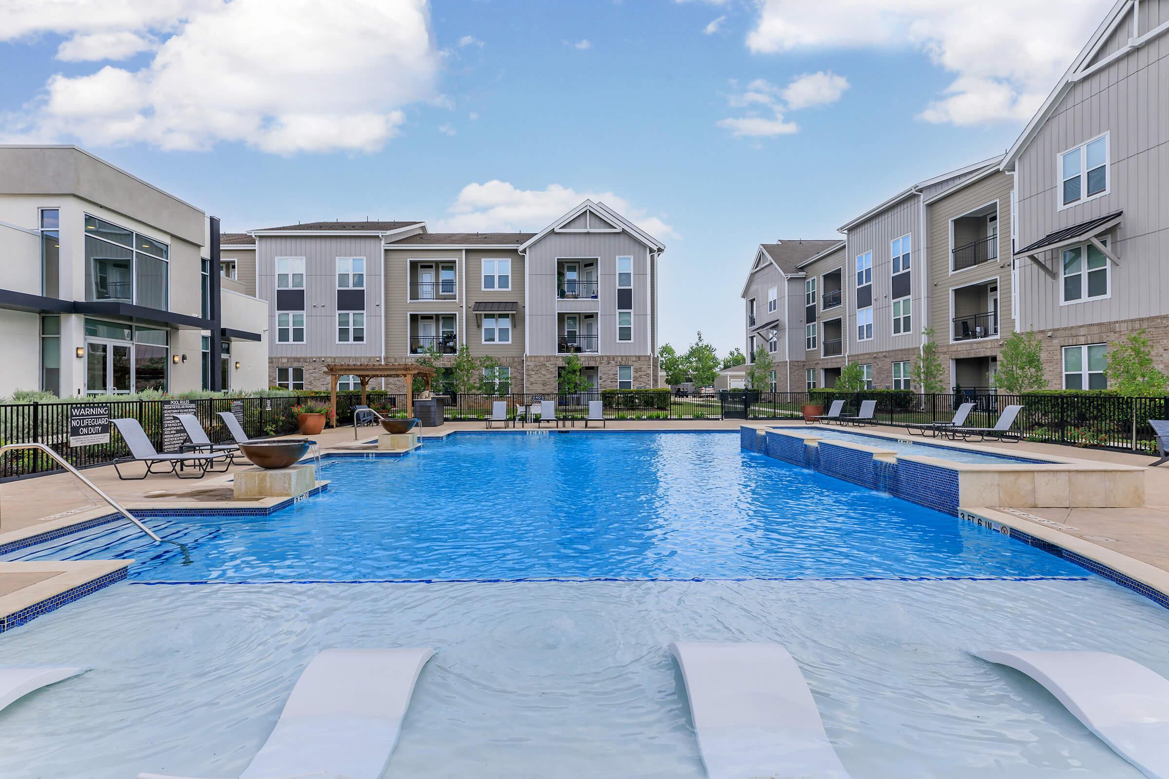 A brightly lit modern apartment complex with a large swimming pool in the foreground. The pool features lounge chairs and a shallow area, surrounded by landscaped greenery. Several buildings of the complex are visible in the background, showcasing contemporary architecture against a blue sky with scattered clouds.