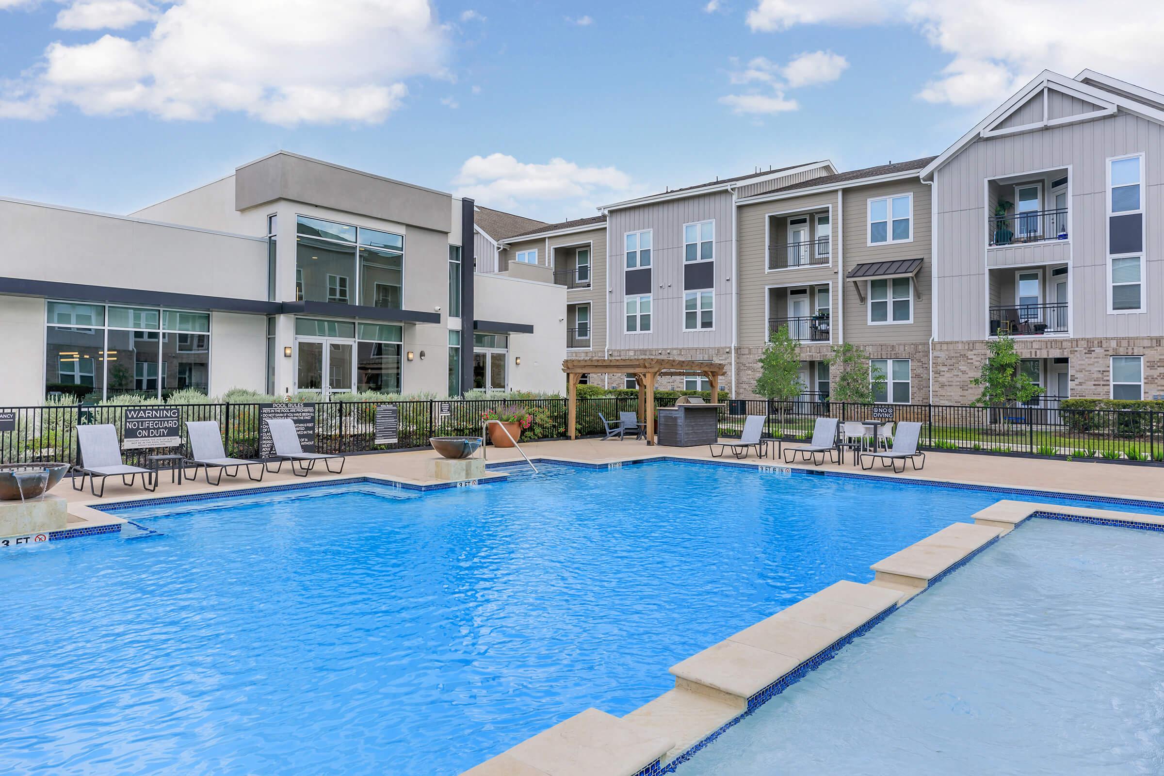 A view of a modern apartment complex featuring a large outdoor swimming pool with lounge chairs and umbrellas. Surrounding the pool are landscaped areas with greenery. The building has a contemporary design with a mix of stone and siding, and large windows reflecting the sky.