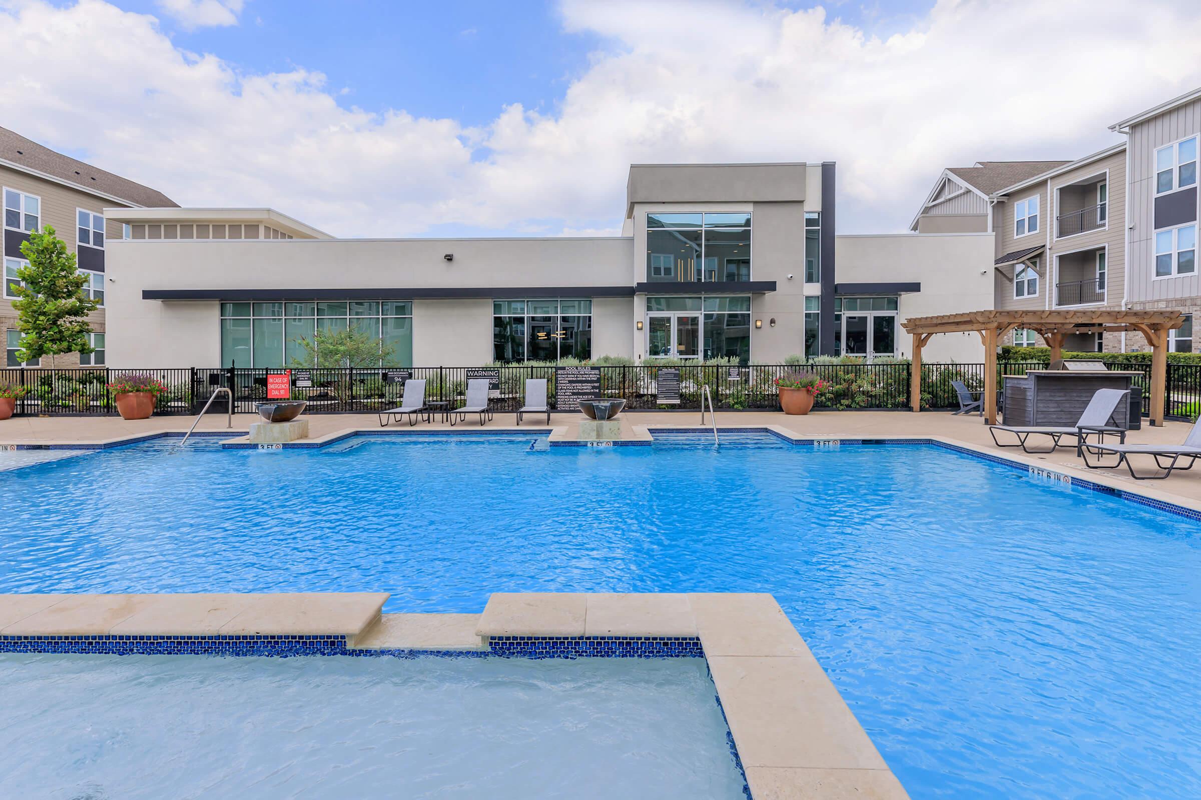 A large outdoor swimming pool with lounge chairs surrounding it. In the background, a modern building features large windows and greenery. The sky is partly cloudy, enhancing the inviting atmosphere of the pool area.