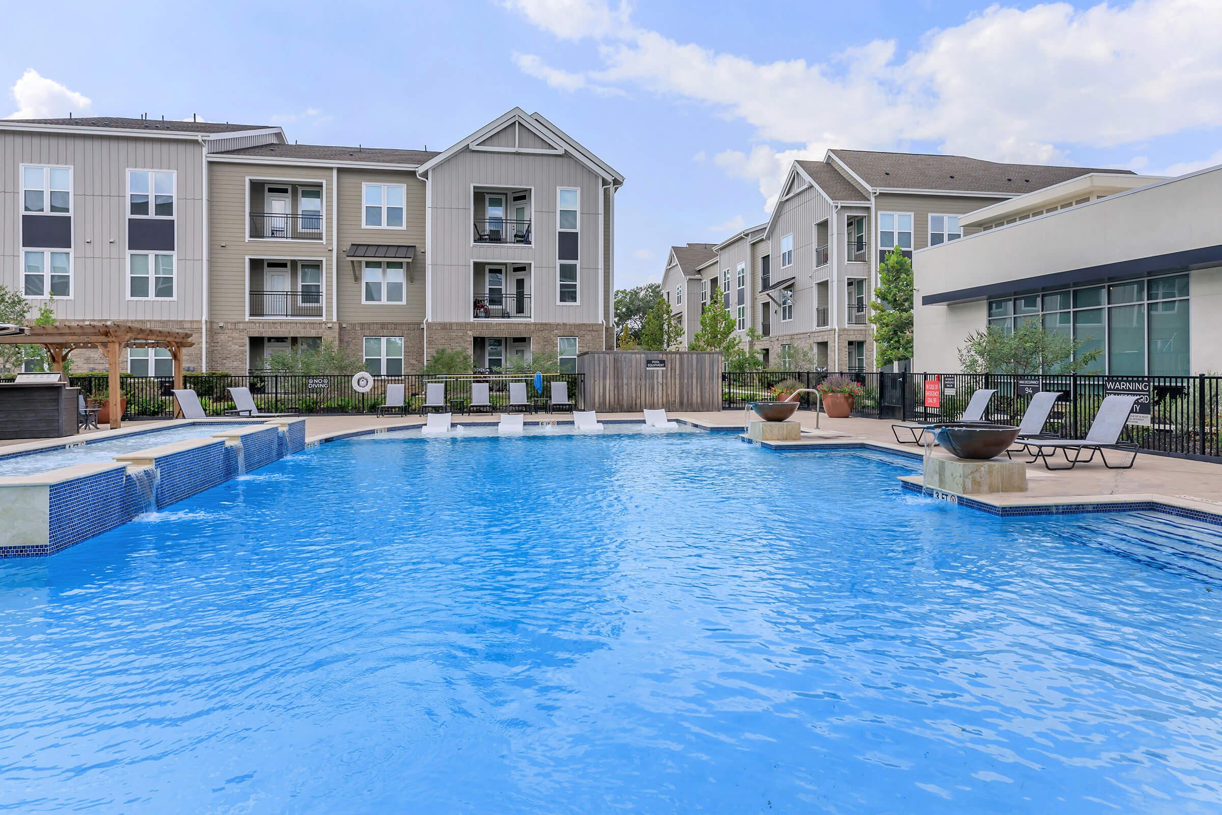 A large, inviting swimming pool surrounded by lounge chairs and modern apartment buildings. The pool features a shallow area and a deep end, with the sky reflecting on the clear water. Nearby, there are outdoor seating areas and greenery, creating a relaxing environment for residents.