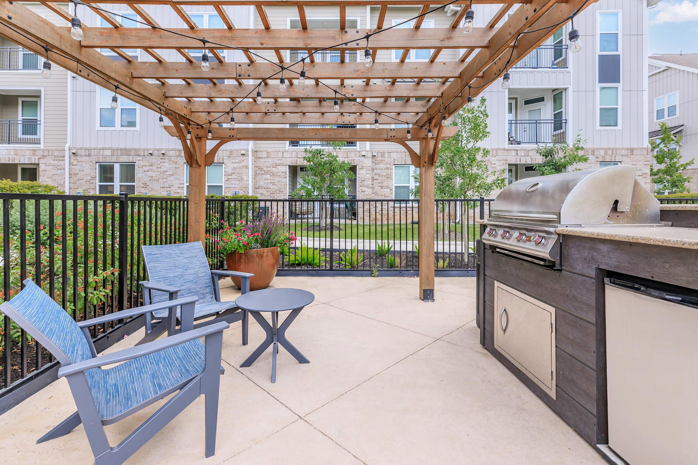 Outdoor grilling area featuring a wooden pergola, two blue chairs, and a small round table. A gas grill is positioned on a stone countertop, surrounded by greenery and a black railing. Soft string lights hang above, creating a cozy atmosphere in a modern residential setting.
