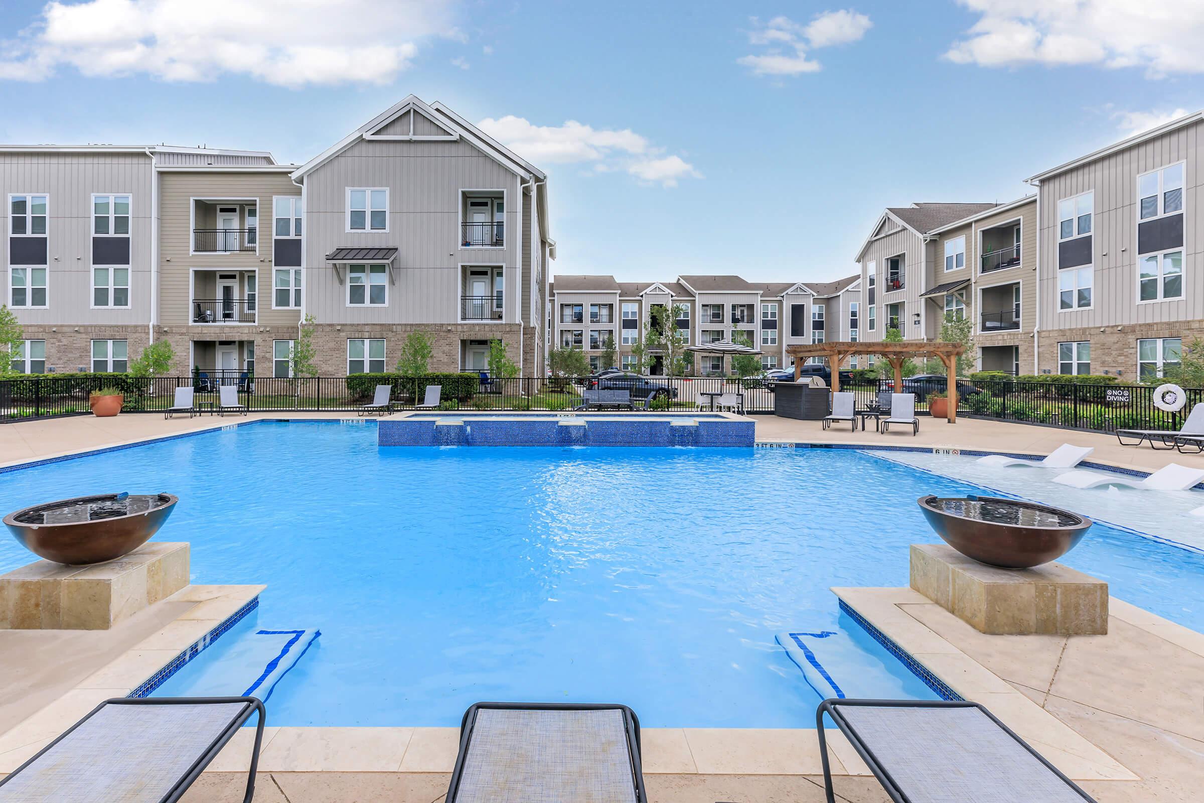 A resort-style swimming pool surrounded by lounge chairs, potted plants, and a stone patio, set in front of modern apartment buildings with large windows. The pool features a tiled edge and a serene atmosphere with clear blue water under a bright sky.