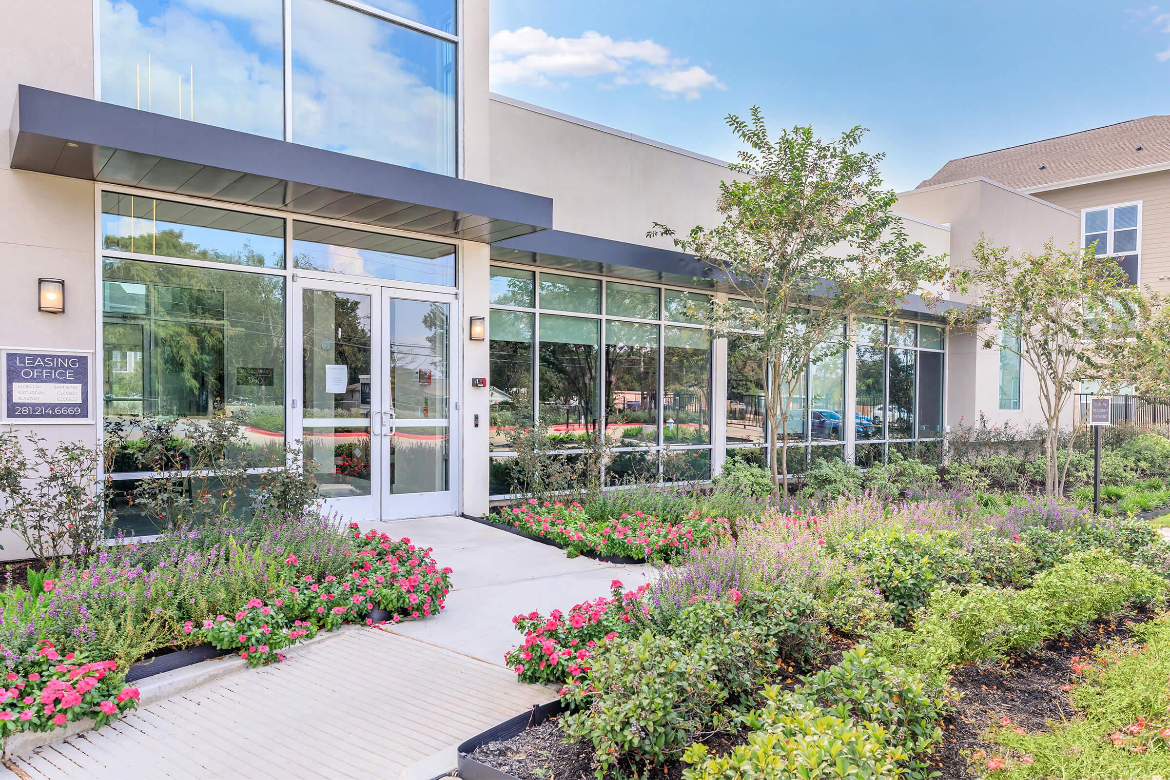 A modern leasing office with large glass windows, surrounded by colorful flower beds and neatly trimmed shrubs. The entrance features a welcoming path leading to the door, and the building is set against a clear blue sky.