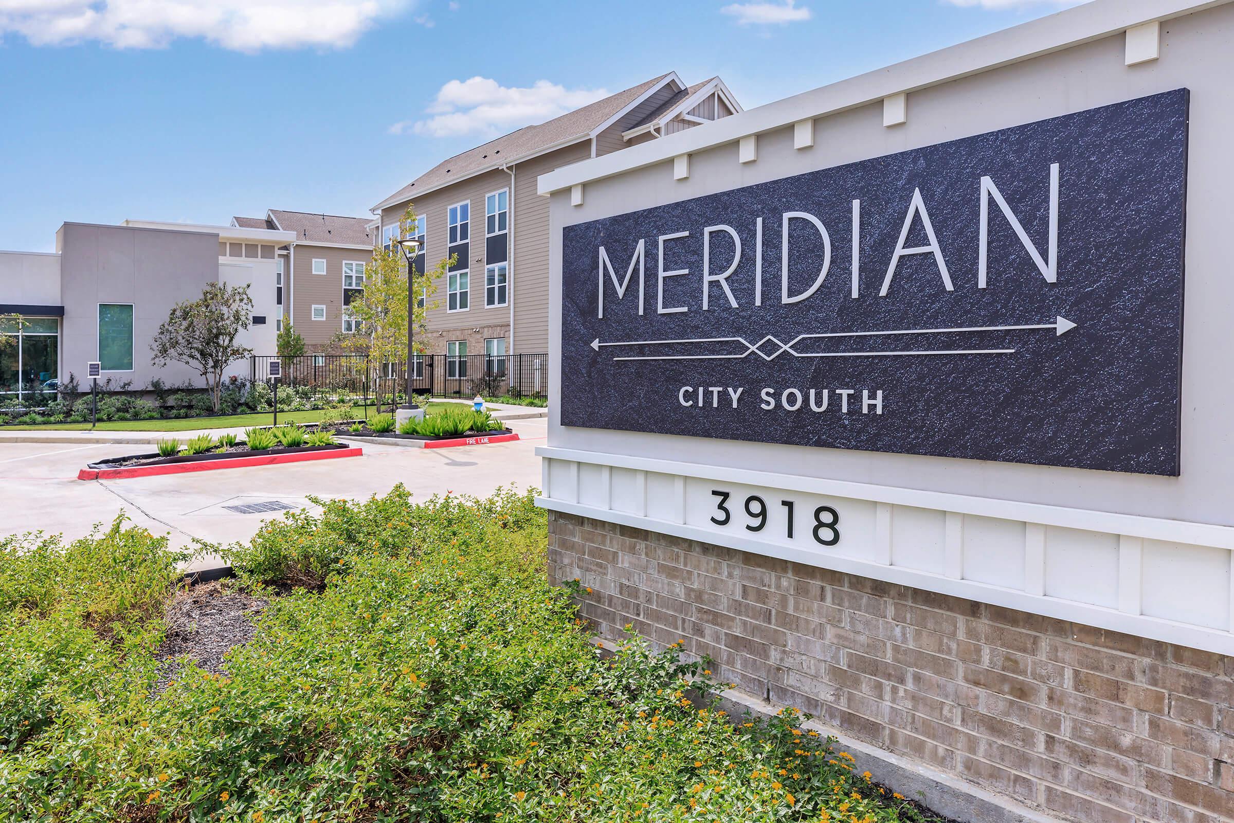 A modern sign for "Meridian City South" featuring the name prominently displayed in white against a dark background, surrounded by landscaped greenery. In the background, there are contemporary apartment buildings under a clear blue sky.