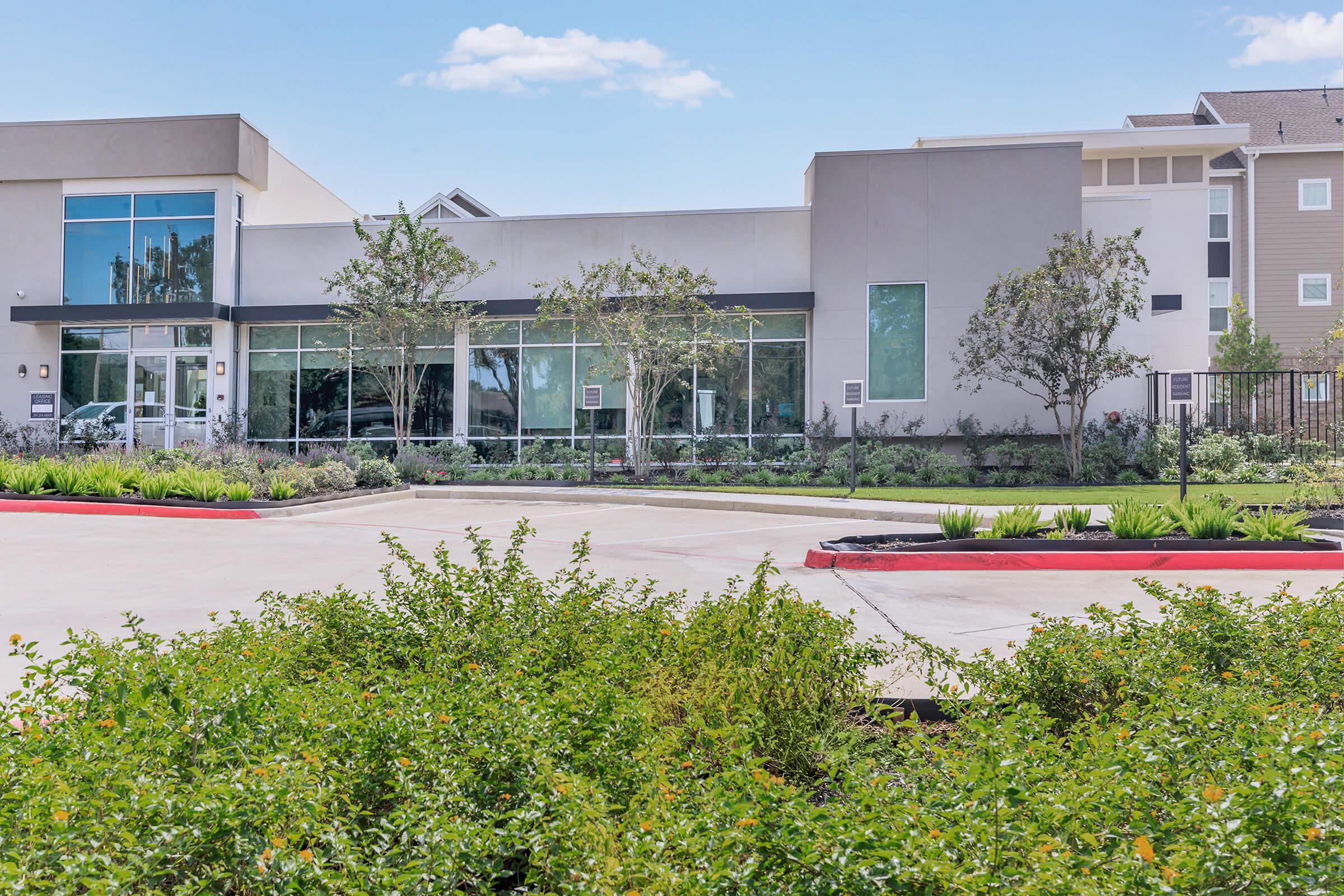 Modern building with large windows and a landscaped parking area. The front features greenery, including small trees and shrubs, while a smooth driveway leads to the entrance. The sky is clear with a few clouds, and the overall setting appears inviting and well-maintained.