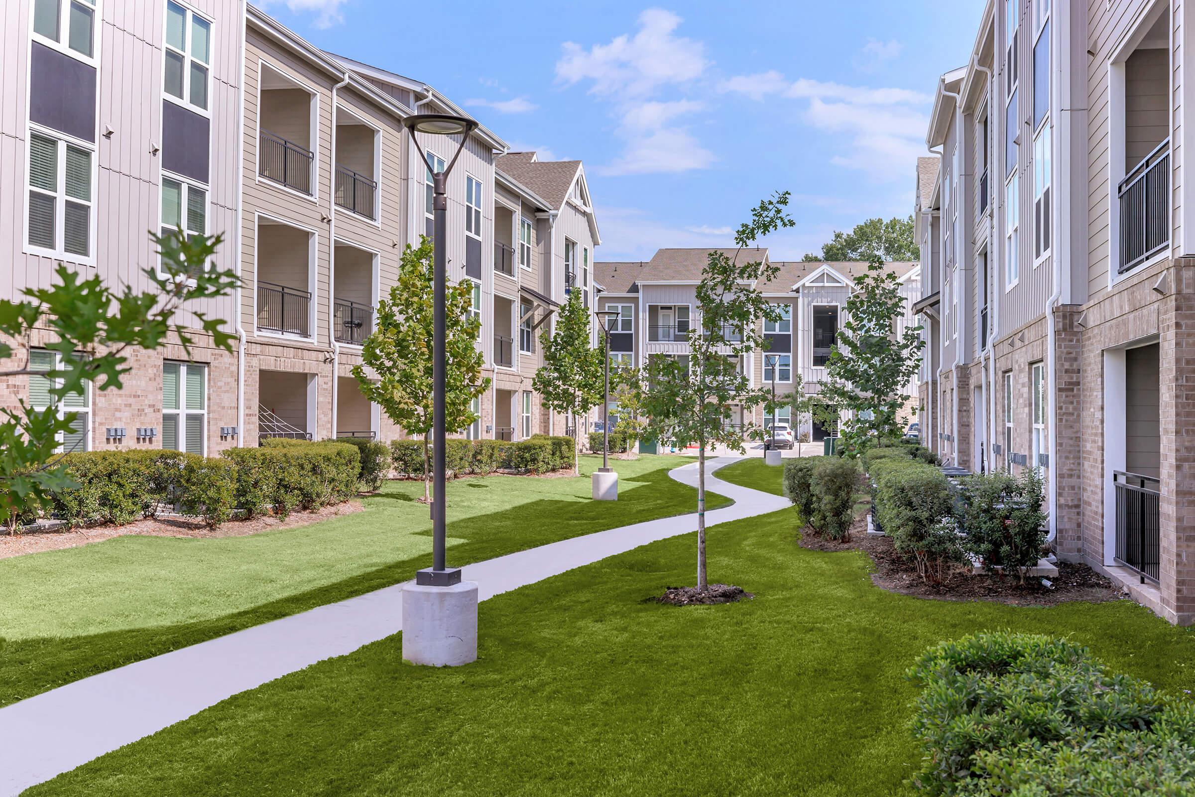 A landscaped pathway winding through an apartment complex, flanked by neatly trimmed bushes and small trees. Light-colored buildings with balconies and large windows are visible in the background under a blue sky with a few clouds. The scene conveys a peaceful, residential atmosphere.