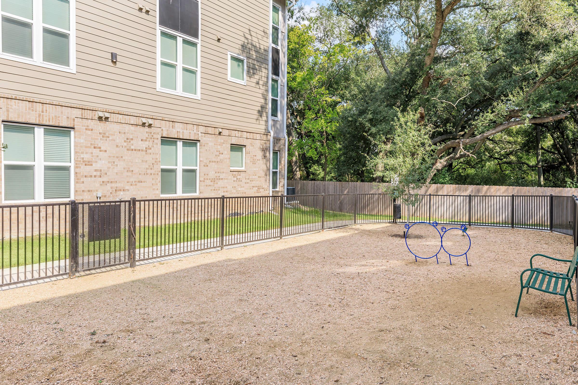 A fenced playground area with a gravel surface, featuring a pair of blue circular play structures and a green bench. The background shows a modern building with large windows and greenery around it.