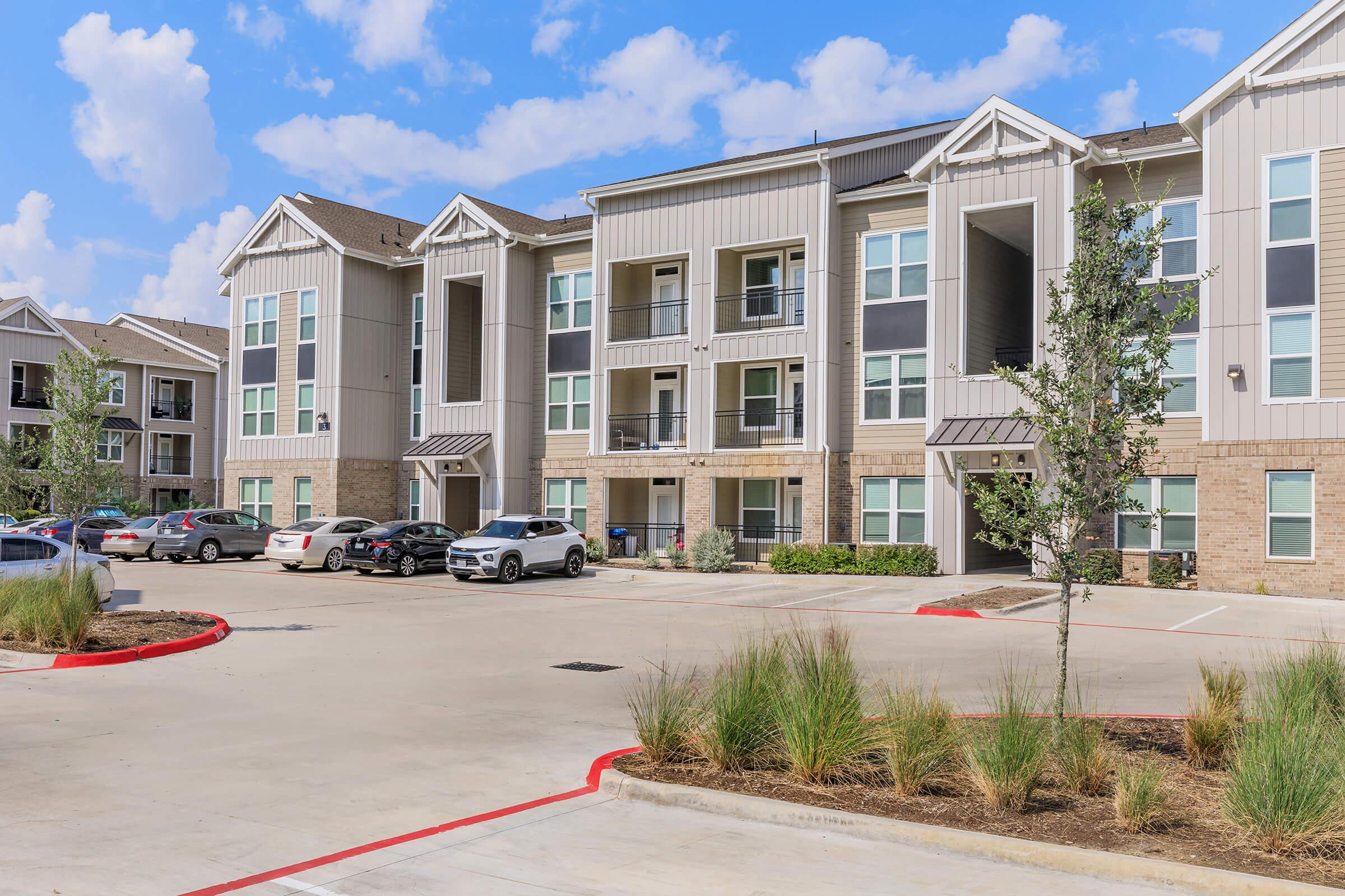 A modern apartment complex featuring multiple three-story buildings with balconies, surrounded by a paved parking lot. Green landscaping with shrubs and small trees enhances the area. Blue skies with scattered clouds create a bright atmosphere.