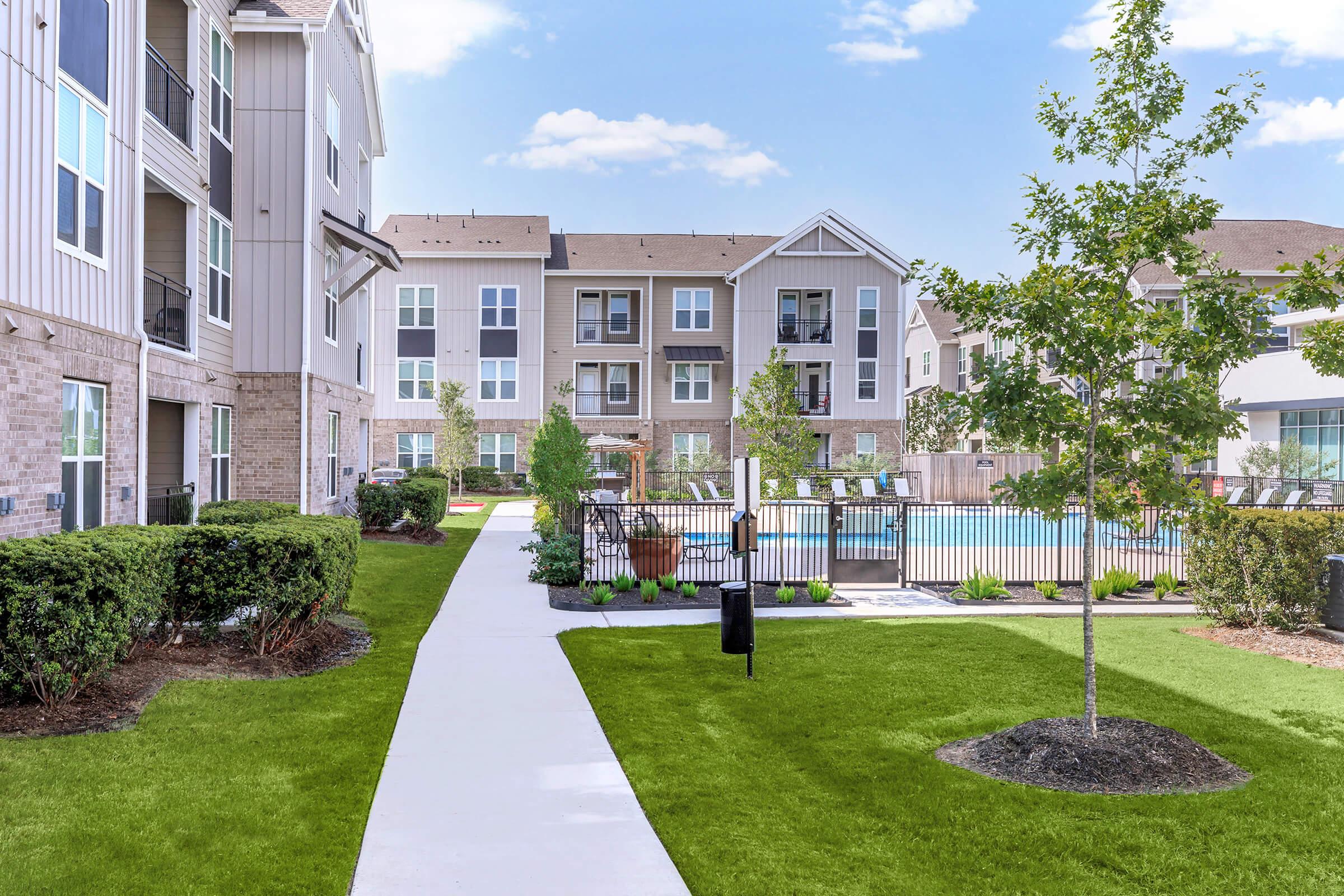 A well-maintained apartment complex featuring a landscaped courtyard with green grass, walking paths, and trees. In the background, there is a swimming pool surrounded by lounge chairs, with several multi-story buildings visible. The sky is clear and bright.
