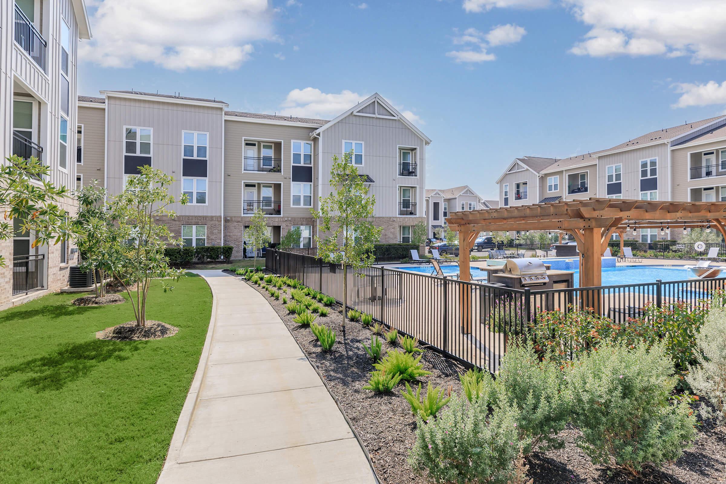 A well-maintained apartment complex with modern buildings, a spacious sidewalk, and landscaped gardens. A swimming pool is visible in the background, surrounded by lounge chairs and a pergola. Lush greenery and small trees line the path, providing a welcoming and serene outdoor environment.