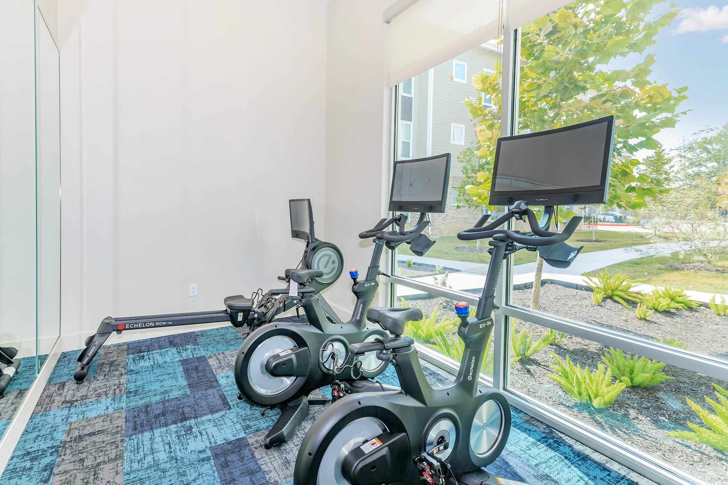 A bright fitness room featuring two stationary bikes with screens, positioned near a large window with a view of greenery outside. The floor is covered with blue carpet tiles, creating a modern and inviting atmosphere for exercise.