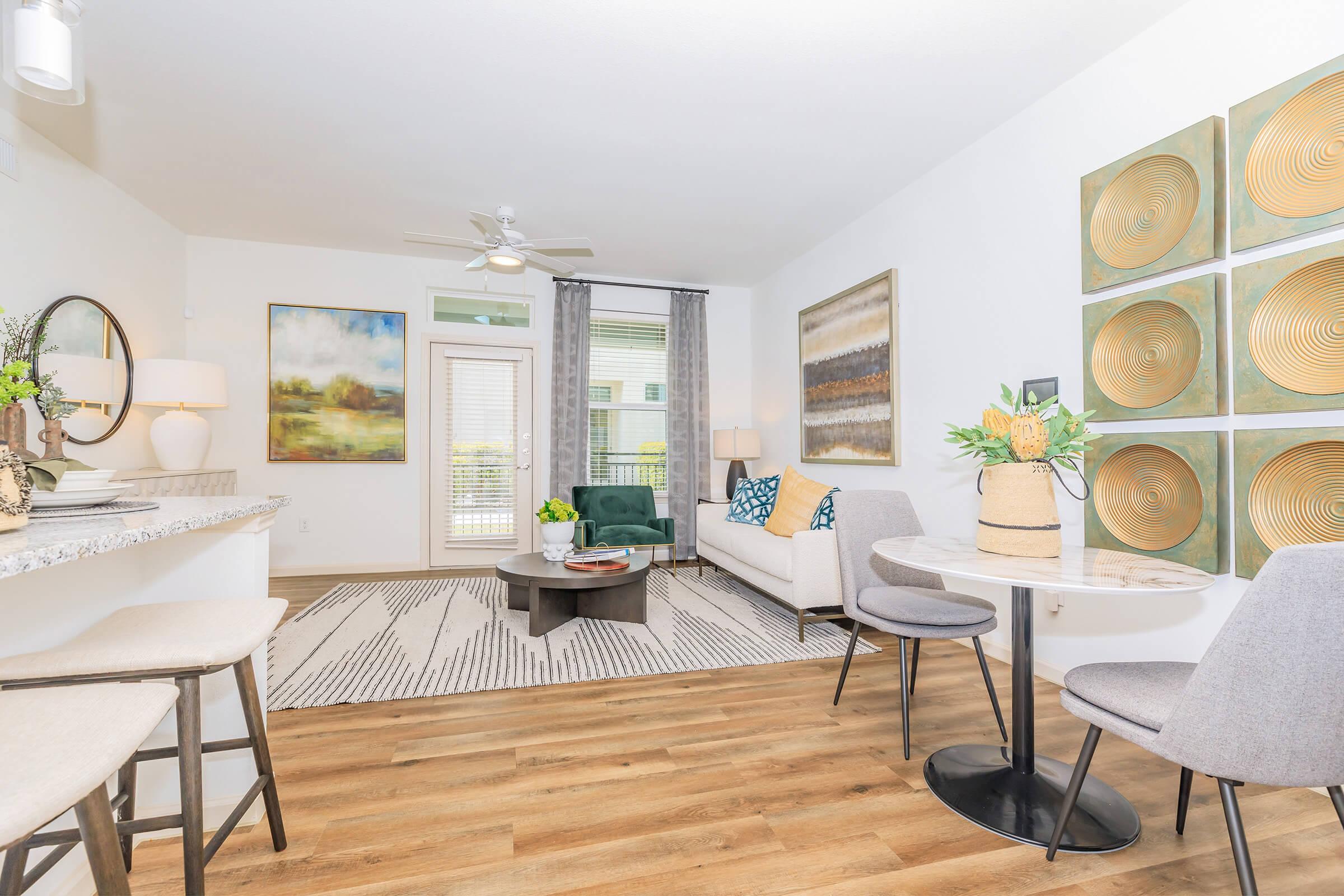 A modern living area featuring a light-colored couch with blue and beige accents, a round coffee table, and stylish decorative art on the walls. There are two dining chairs and a small table, with a textured area rug on the wooden floor. Natural light streams through a sliding glass door, enhancing the airy atmosphere.