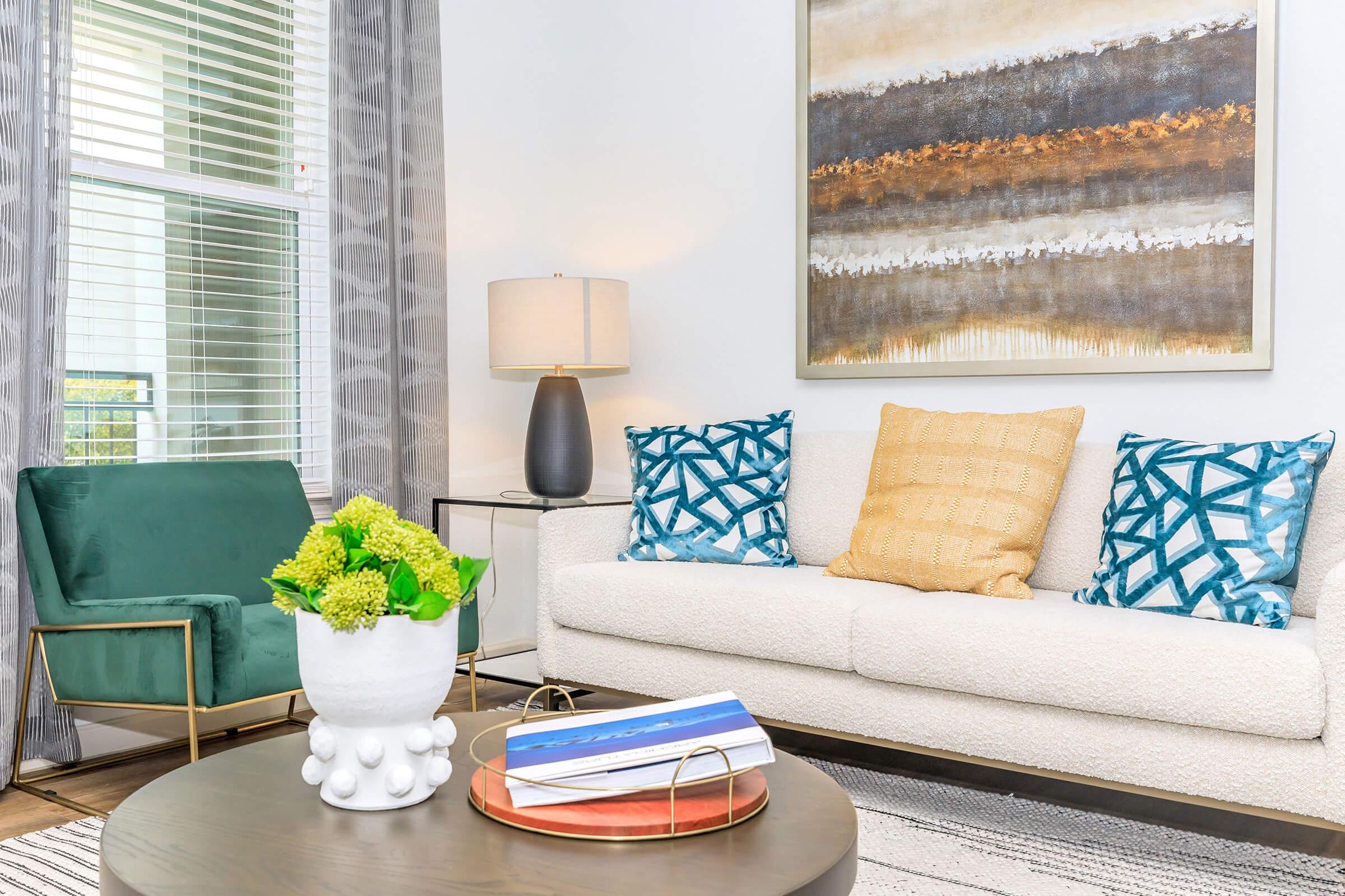 A stylish living room featuring a cozy white sofa adorned with patterned cushions, an elegant green chair, a round coffee table with books and a decorative bowl of flowers, and an illuminated lamp beside a window with patterned curtains. A large abstract art piece hangs on the wall.