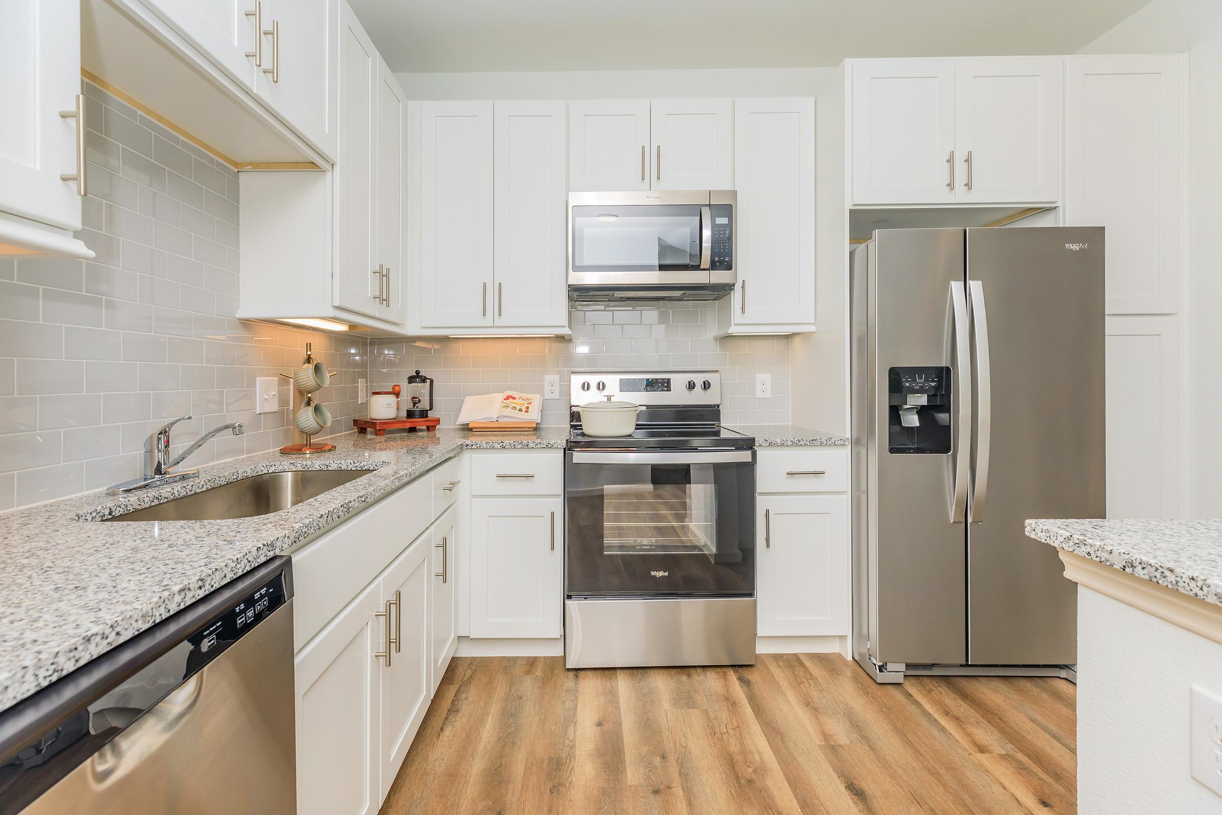 A modern kitchen featuring white cabinets, stainless steel appliances including a microwave, oven, and refrigerator. The countertops are granite, and there is a stainless steel sink. The floor is wooden, and the backsplash includes light gray tiles. Bright and inviting overall.