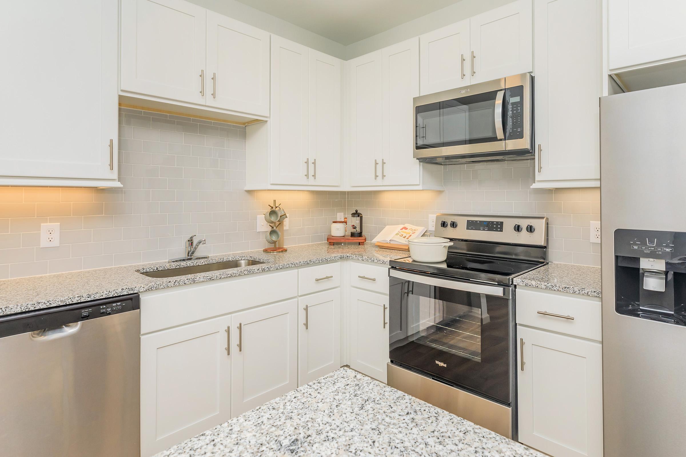 Modern kitchen with white cabinets, granite countertops, and stainless steel appliances. The setup includes an oven, microwave, and a sink, with soft lighting under the cabinets. A decorative plant and kitchen essentials are placed on the counter, contributing to a clean and inviting atmosphere.