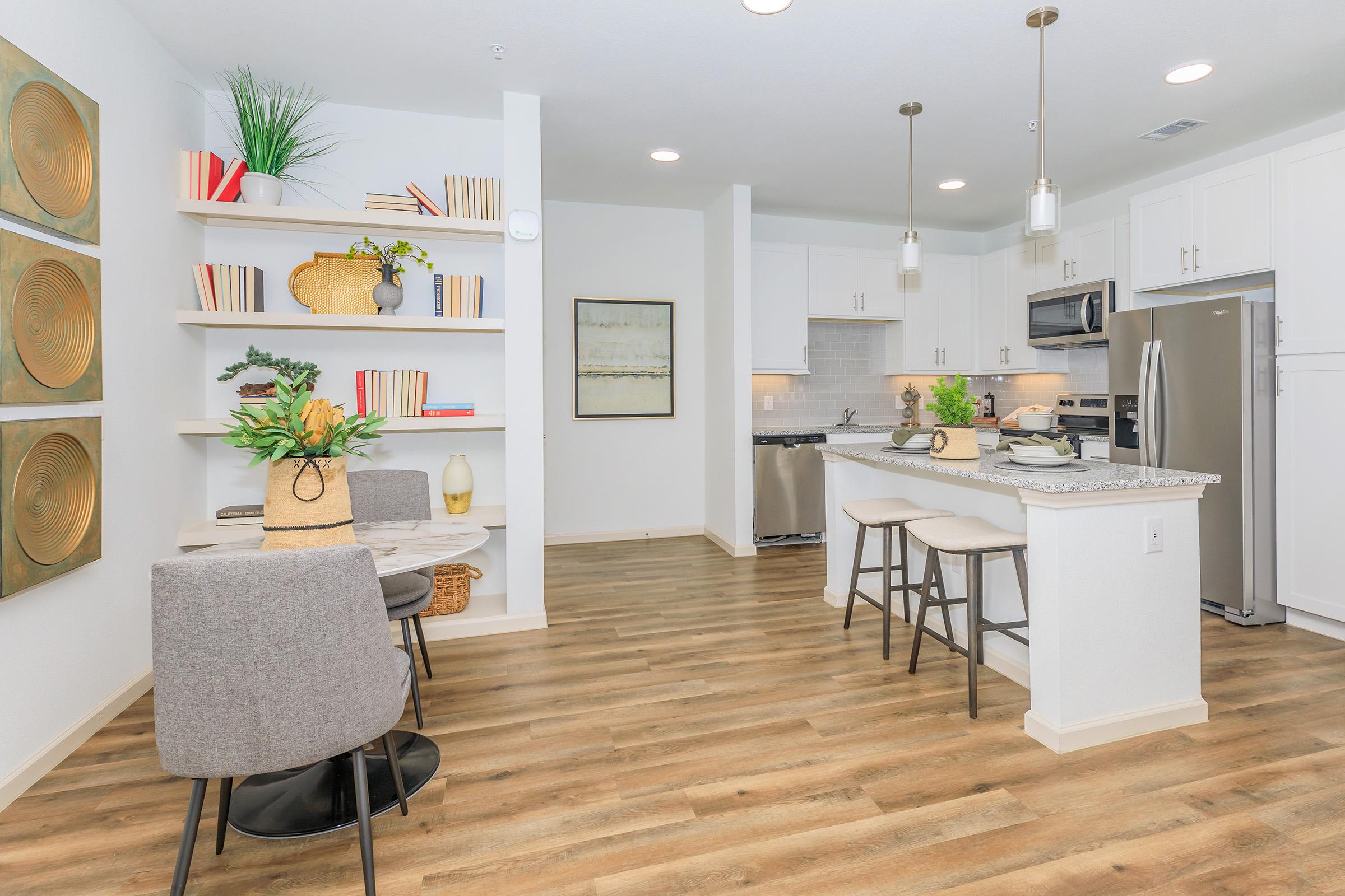 A modern kitchen and dining area featuring a round table with two chairs, a kitchen island with bar stools, and white cabinetry. Decor includes plants, framed artwork, and neatly arranged books on shelves, with warm wooden flooring enhancing the contemporary style.