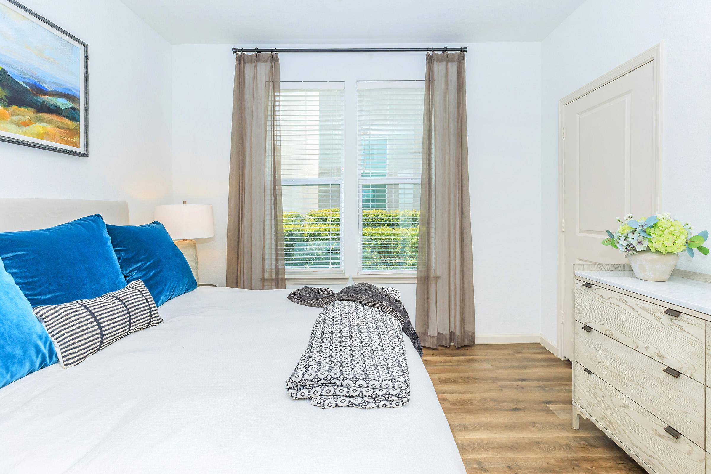 A cozy bedroom featuring a comfortable bed with blue and white pillows, a patterned throw blanket, a light wood nightstand, and a window with sheer curtains allowing natural light. A painting hangs above the bed, and a decorative plant adds a touch of greenery to the space.