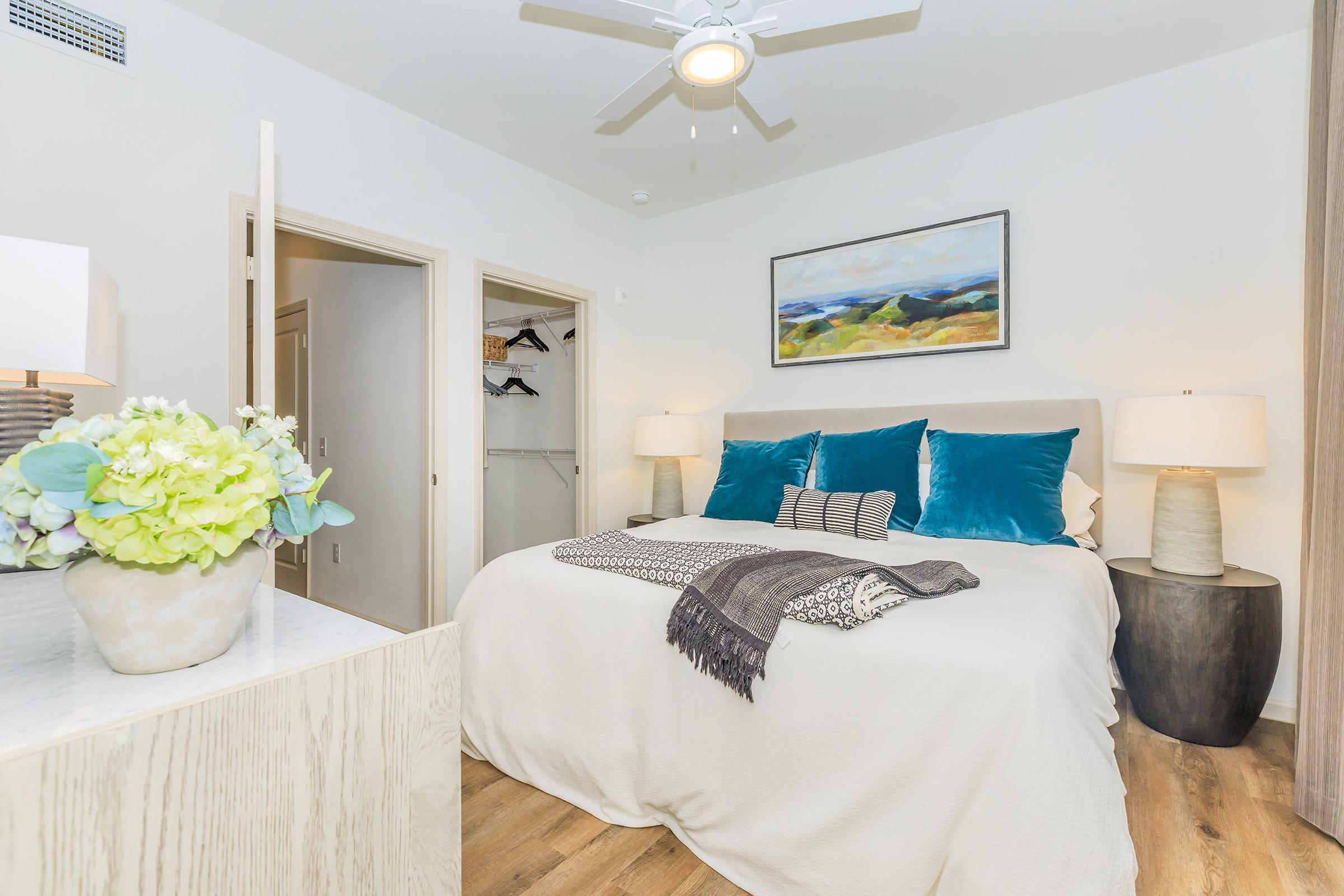 A cozy bedroom featuring a large bed with plush pillows and a textured throw blanket. There are two stylish lamps on either side of the bed, and a decorative plant on the dresser. The walls are light-colored, and a framed landscape painting adds a touch of color. A closet with hanging space is visible in the background.