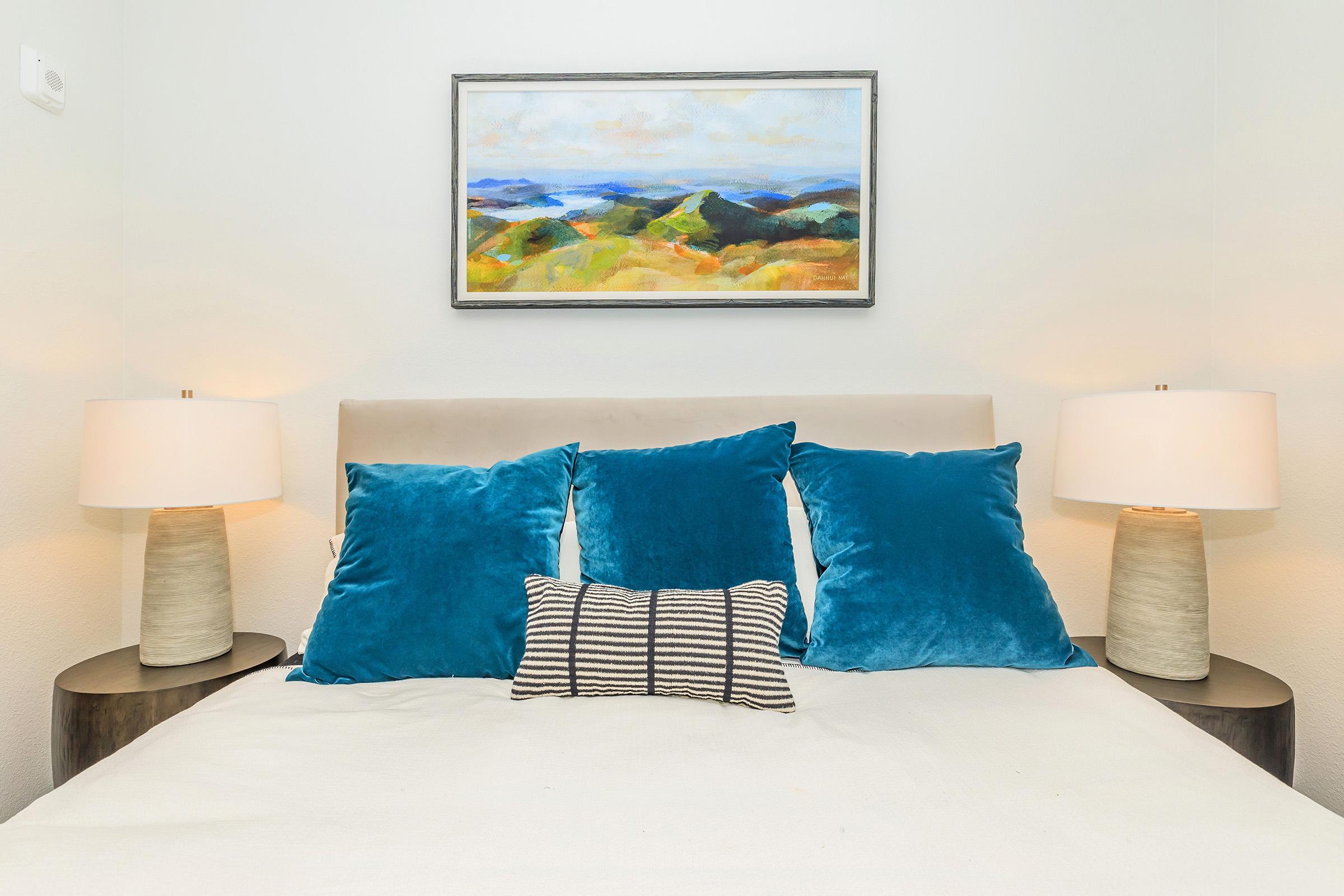 A cozy bedroom featuring a bed with a light-colored headboard adorned with four blue cushions and a striped decorative pillow. Two elegant lamps with textured bases are placed on either side of the bed. Above the headboard, a landscape painting in soft colors adds a touch of art to the serene atmosphere.