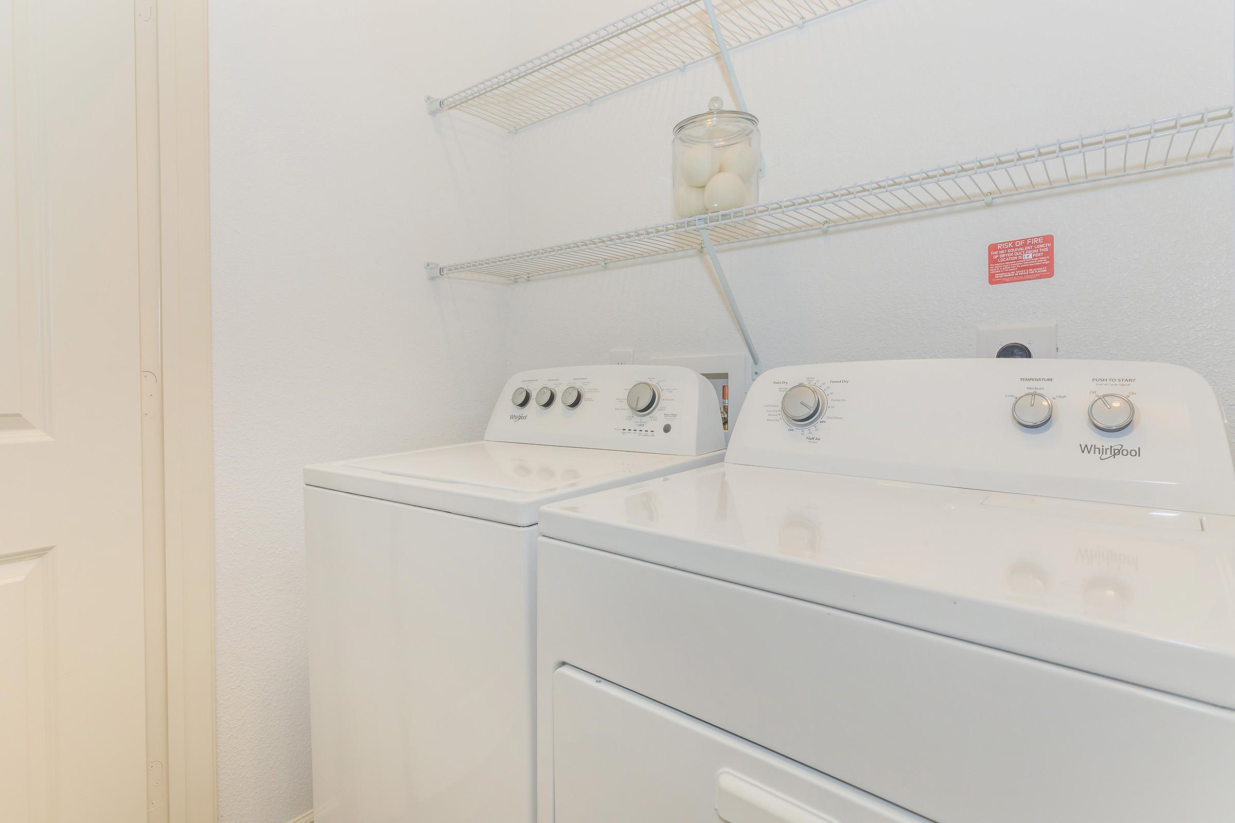 A clean laundry room featuring a white Whirlpool washing machine and dryer side by side. Above them, a shelf holds a clear jar with eggs. The wall is painted white, and there is a door partially visible on the left. The space appears neat and well-organized.
