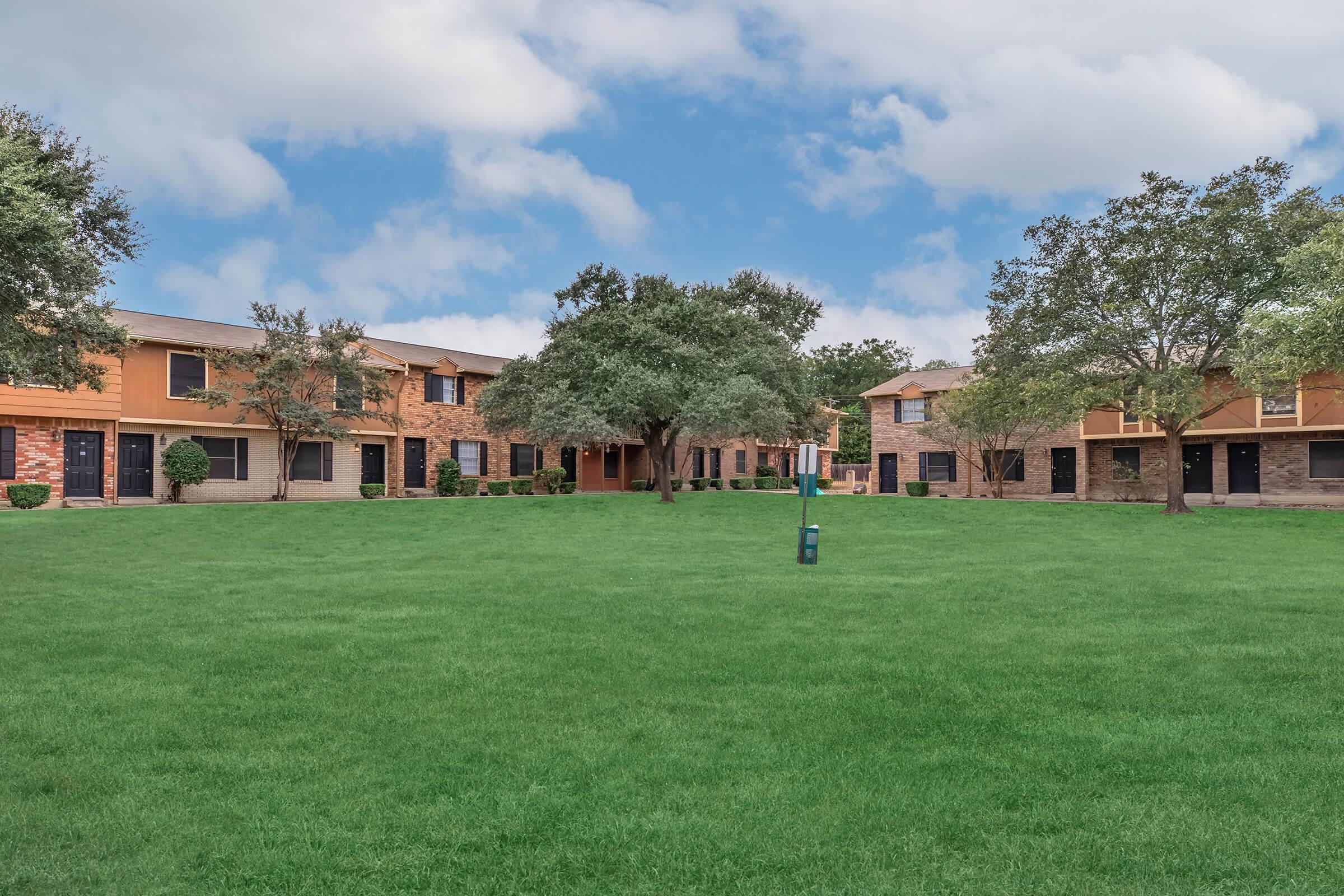 A residential apartment complex featuring two-story buildings arranged around a spacious green lawn, with trees providing shade. The sky is partly cloudy, creating a serene atmosphere. The buildings have a mix of stone and siding exteriors, with a walkway leading through the lawn.