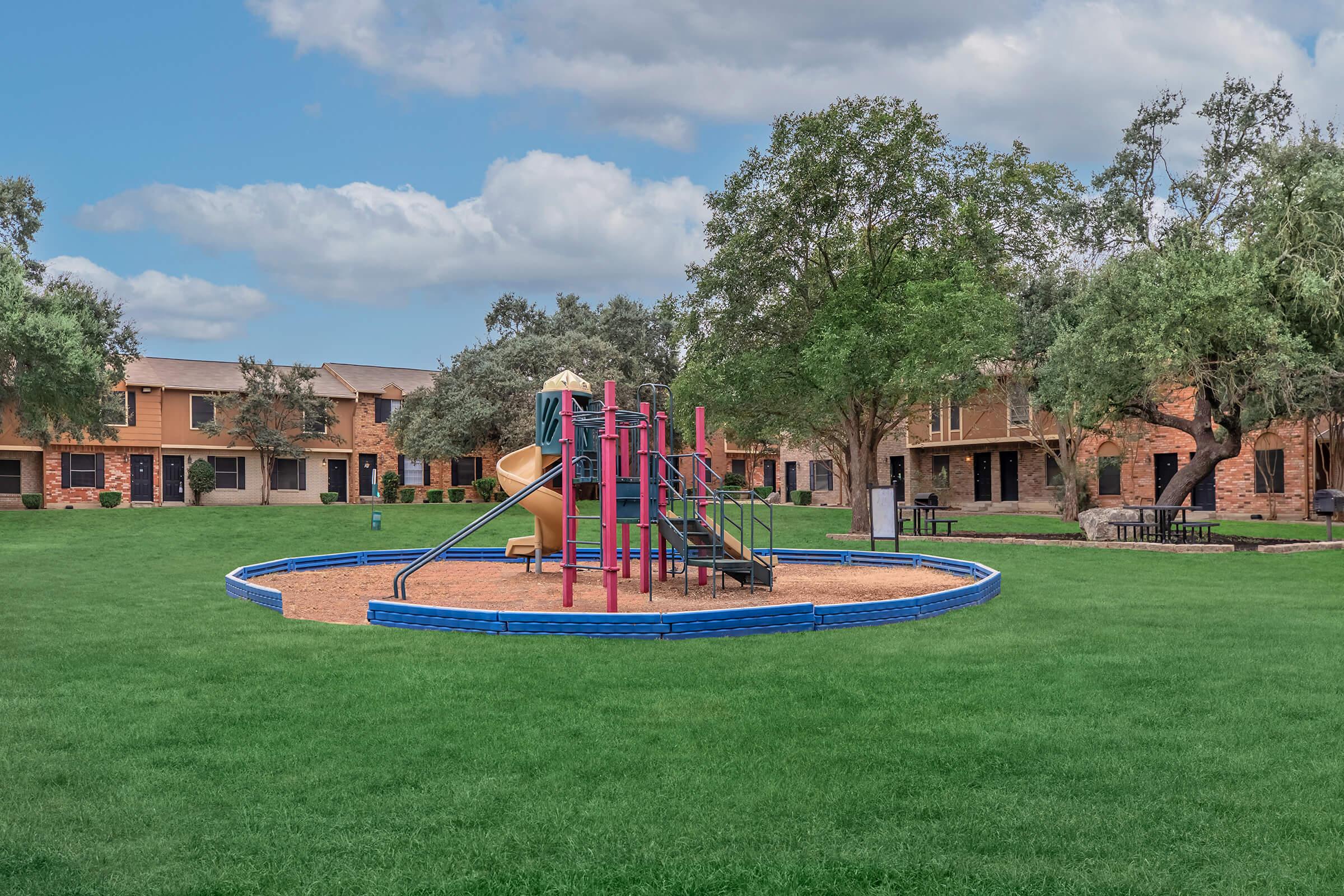 A playground featuring a slide and climbing structure surrounded by a soft, sandy area. The playground is situated in a green lawn area, with several trees and residential buildings in the background under a partly cloudy sky. Bright colors and open space create a welcoming environment for children.