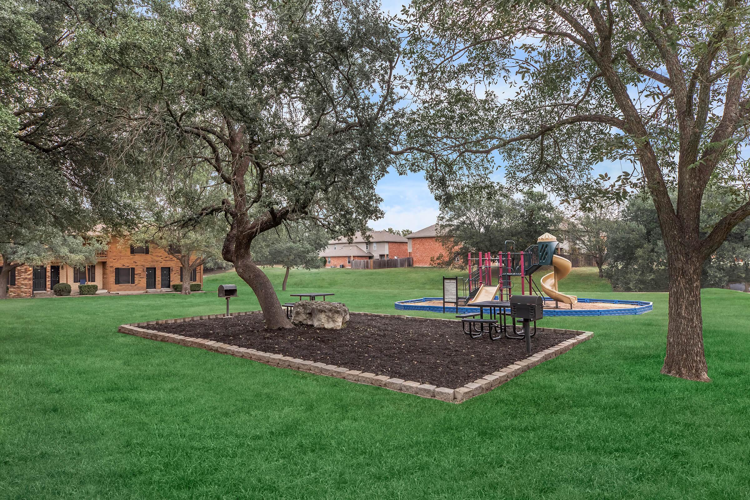 A green park area featuring a playground with a slide and climbing structure, surrounded by trees. There are picnic tables and a barbecue grill nearby, set against a backdrop of residential buildings. The ground is covered in grass and mulch.