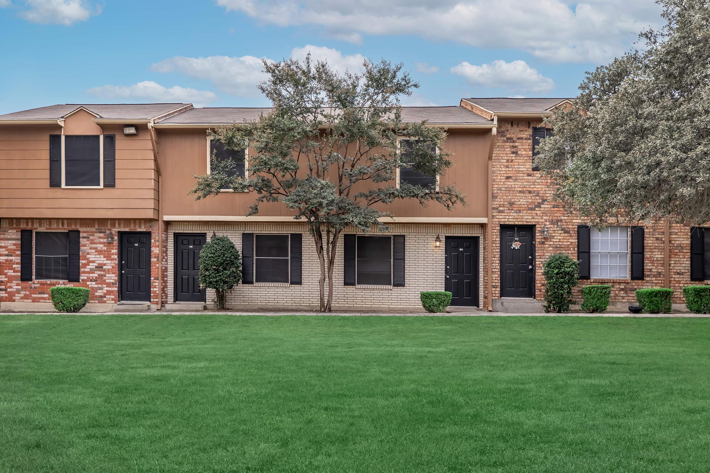 A row of three townhouses featuring a mix of brick and siding exteriors. Each unit has a front door flanked by small shrubs, with a large grassy area in front. The backdrop includes a few trees and a partly cloudy sky, creating a serene suburban atmosphere.