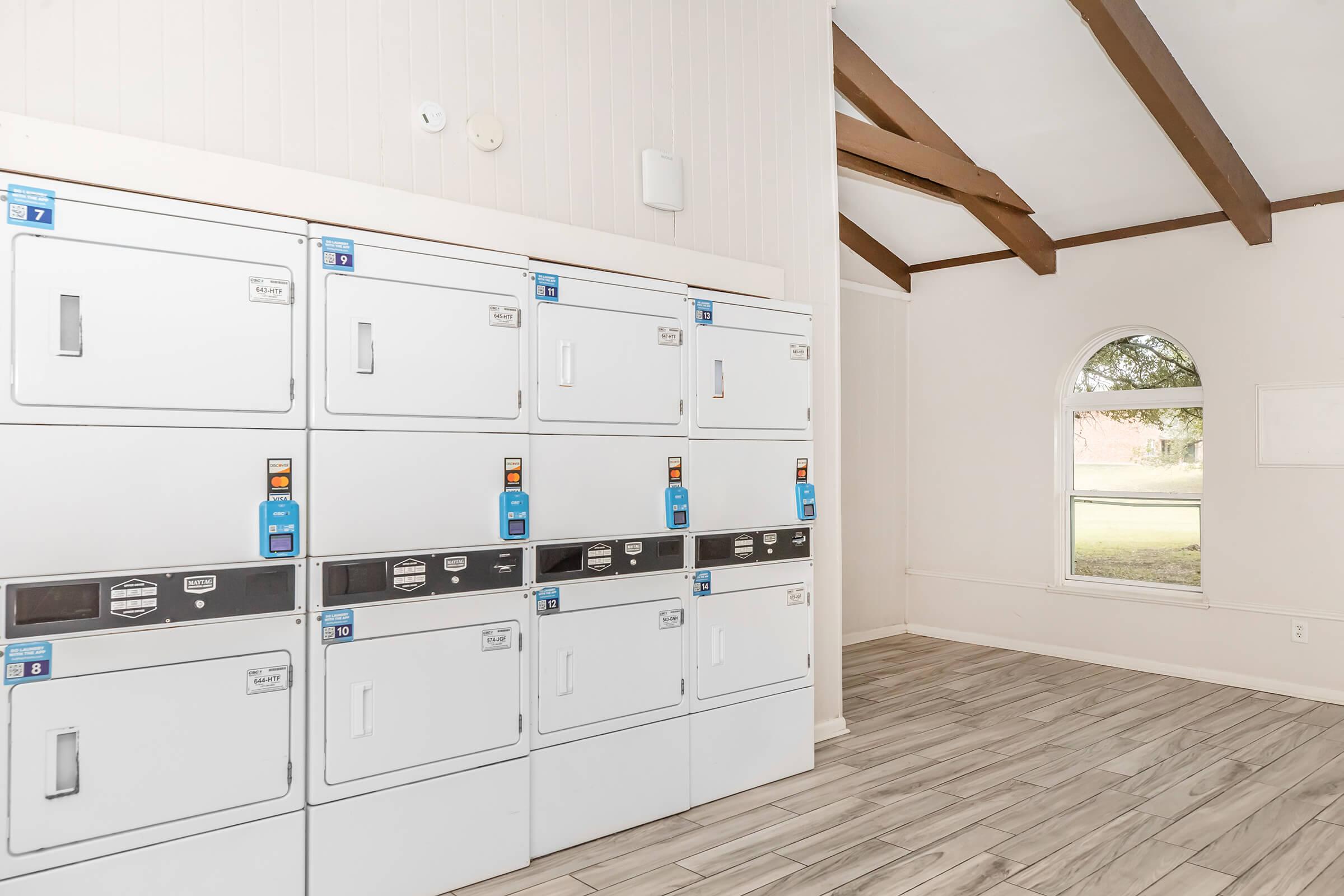A bright, modern laundry room featuring several stacked washing machines and dryers, all in white with blue and orange controls. The room has wooden beams on the ceiling and a large arched window allowing natural light to illuminate the space, complementing the tiled floor.