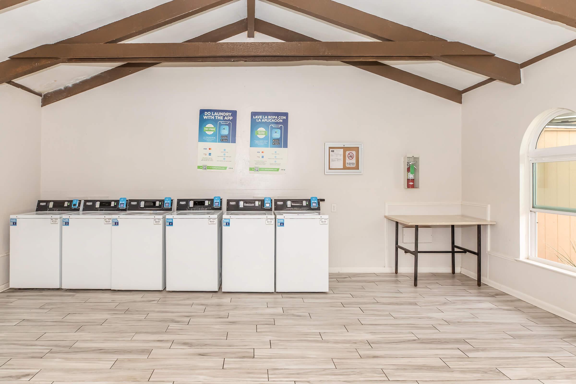 A bright laundry room featuring four white washing machines against a wall with informational posters. The room has a high ceiling with wooden beams and a wooden table in the corner. Natural light enters through a window, illuminating the tiled floor.