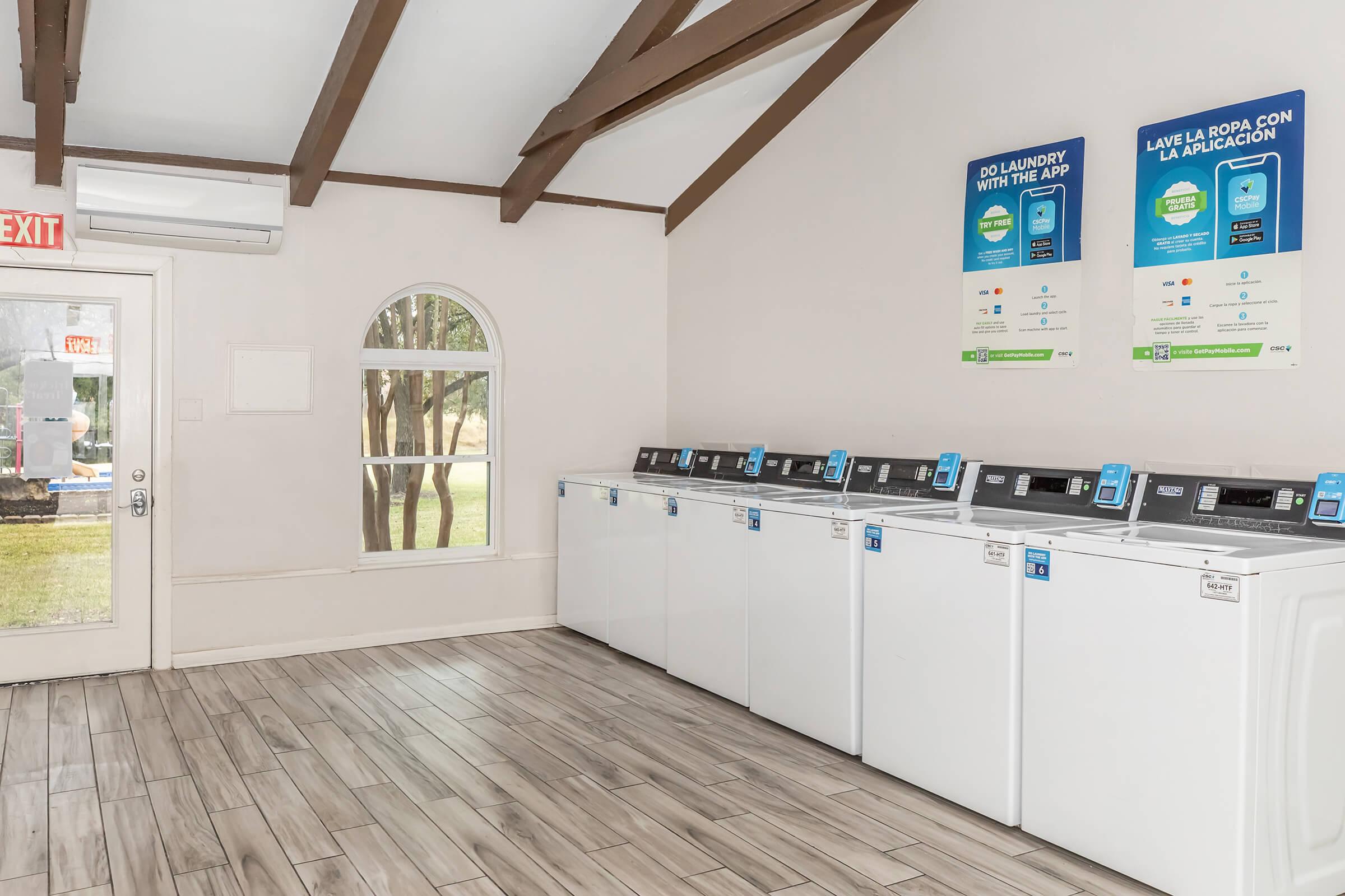 A modern laundry room featuring several white washing machines lined up against the wall, with a large window allowing natural light. Promotional posters are displayed above the machines, and an exit sign is visible. The floor is covered with light-colored tiles, and the ceiling has wooden beams.