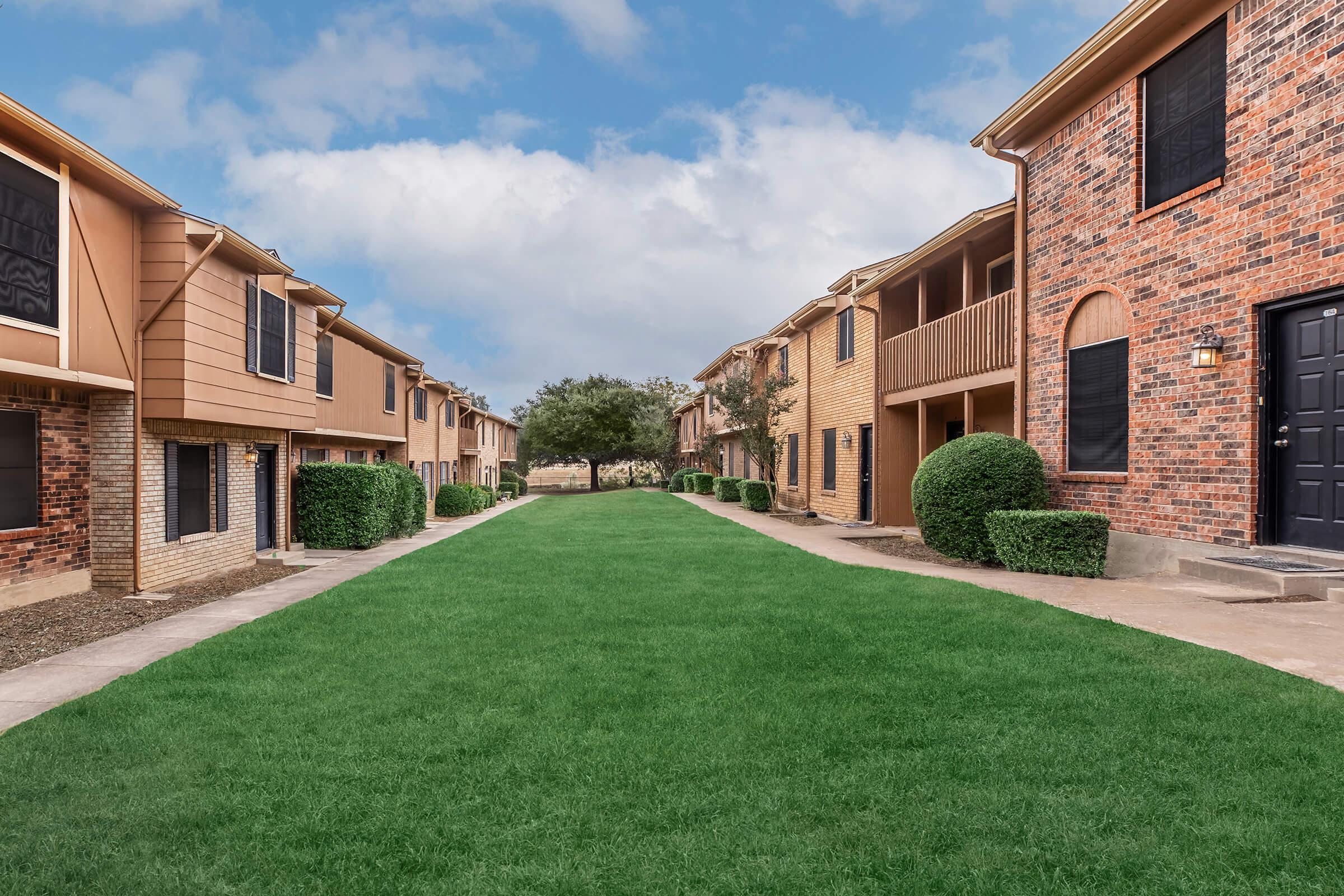 A shared pathway lined with well-maintained grassy areas, leading between two rows of residential buildings. The buildings feature a mix of brick and siding exteriors, with some having balconies. A few trees and shrubs add greenery to the scene, under a cloudy sky.
