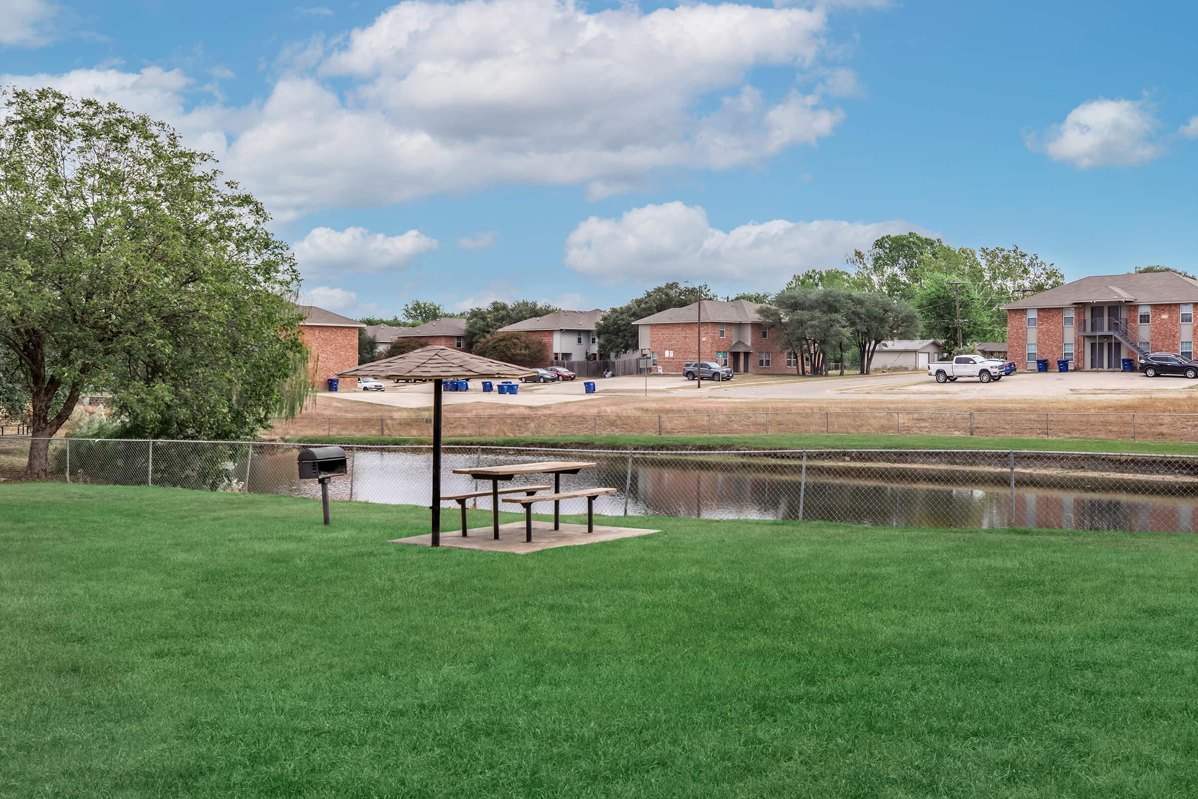 A serene outdoor area featuring a grassy space with a picnic table and grill overlooking a small pond. In the background, there are residential buildings and a tree line under a partly cloudy sky. The scene invites relaxation and outdoor activities.