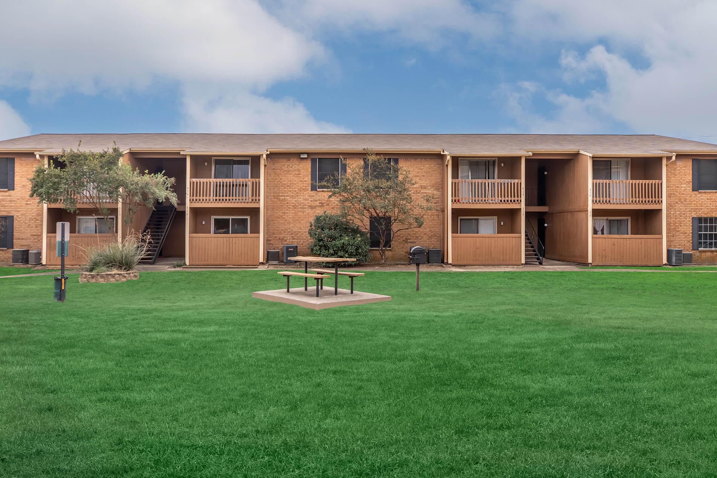 A residential complex featuring two-story brick apartments with balconies. In the foreground, a grassy lawn with a picnic table and grills. The sky is partly cloudy, creating a serene outdoor atmosphere.