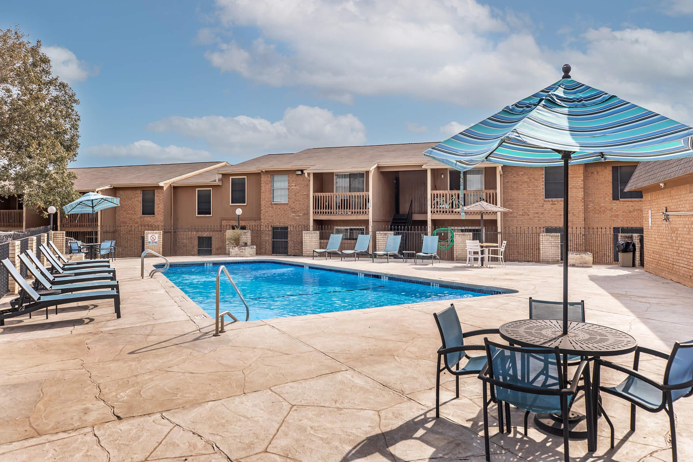 A well-maintained outdoor swimming pool surrounded by lounge chairs and tables. Shade is provided by umbrellas, with two buildings visible in the background. The setting is bright and inviting, ideal for relaxation and leisure activities.