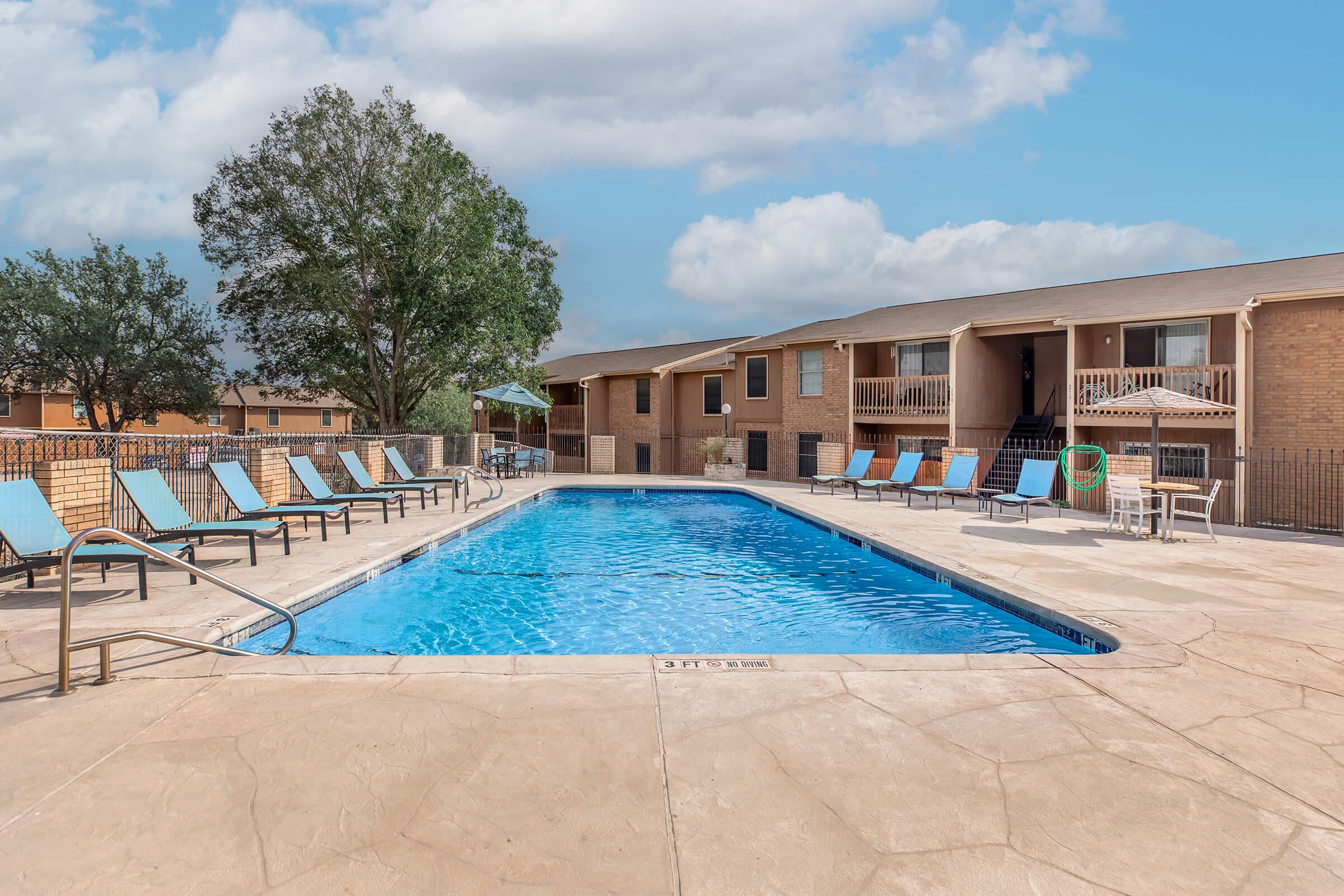 A clear blue swimming pool surrounded by lounge chairs and a patio area. In the background, there are residential buildings and trees under a partly cloudy sky.