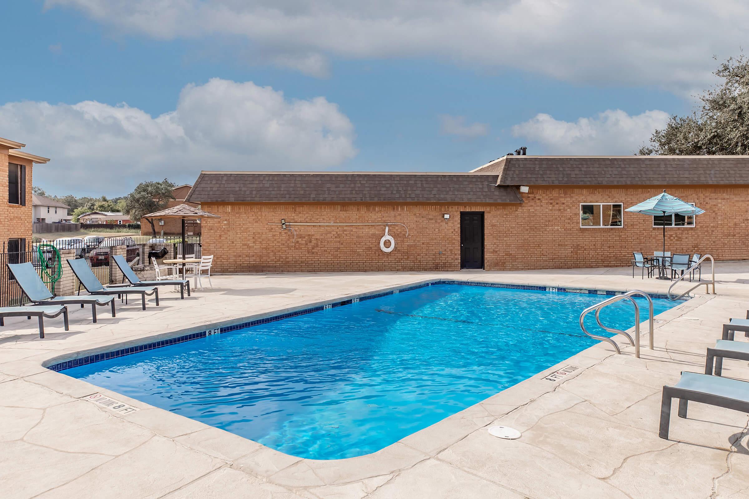 A clean, inviting swimming pool surrounded by a concrete patio, featuring lounge chairs and an umbrella. A brick building is visible in the background under a partly cloudy sky, creating a relaxing atmosphere for leisure and recreation.