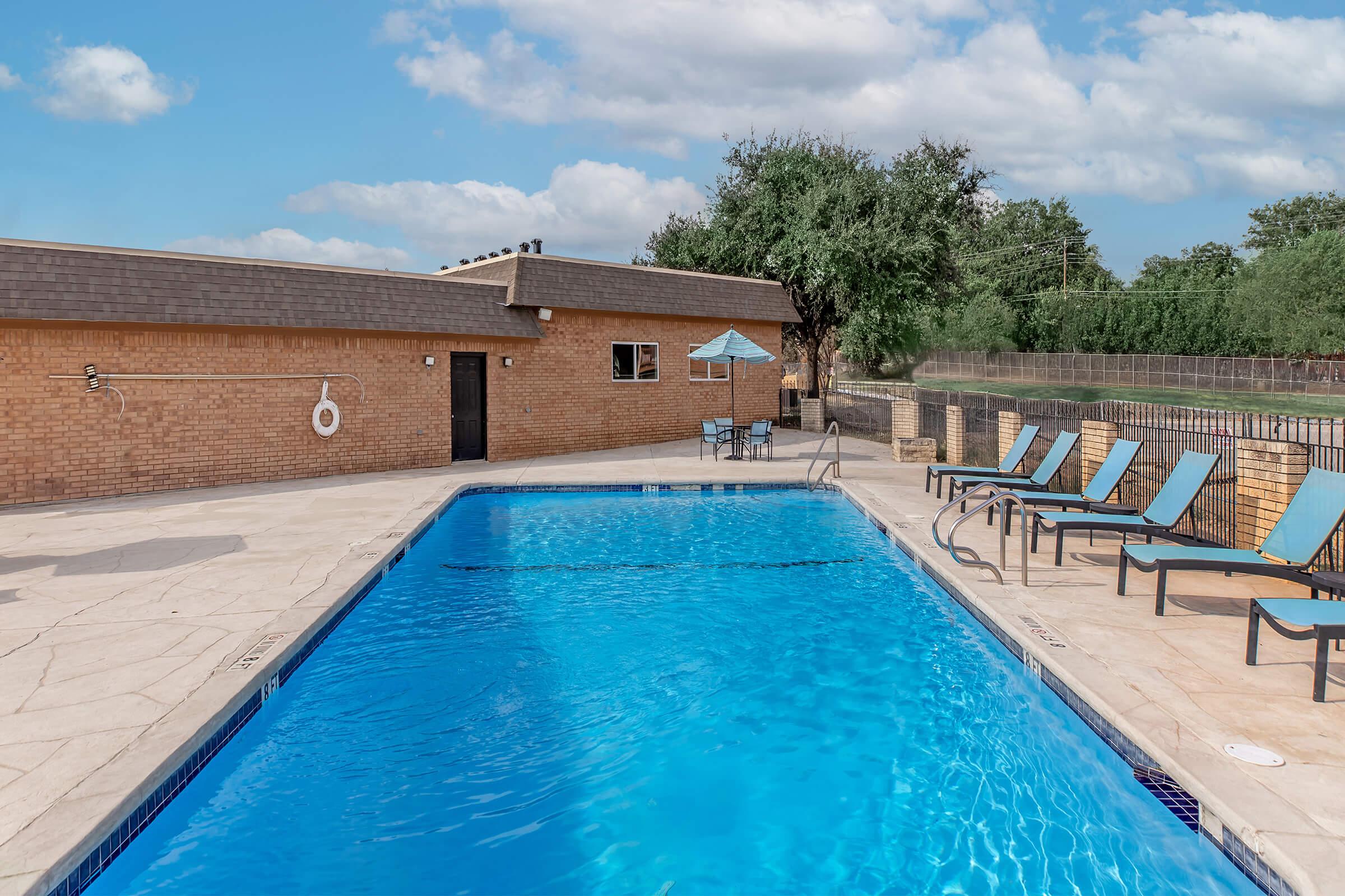 A clear blue swimming pool surrounded by lounge chairs on a tiled deck. There is a shaded table with an umbrella nearby, and a brick building can be seen in the background. Lush greenery and a fence are visible in the distance, under a partly cloudy sky.