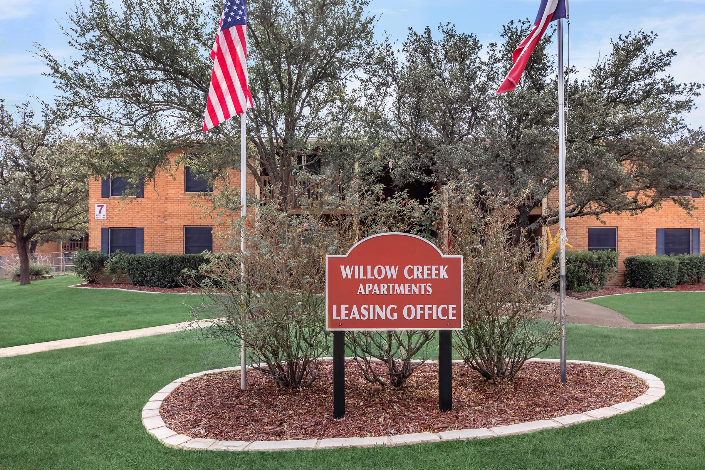 Sign for Willow Creek Apartments leasing office surrounded by landscaped grass and shrubs, with two flags flying in front of a brick building.
