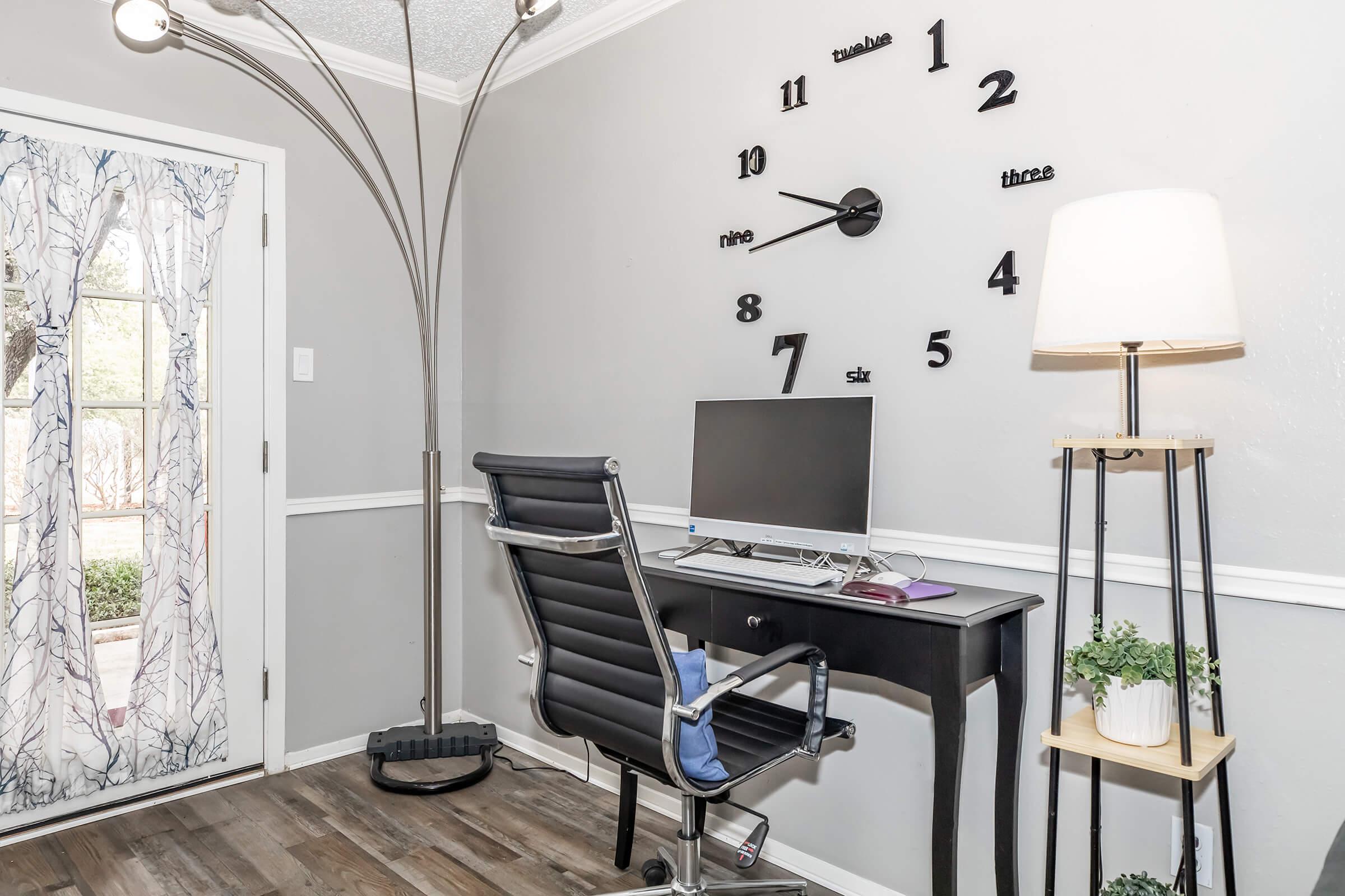 A modern home office space featuring a sleek black desk with a computer monitor, a black office chair, and stylish lighting. The wall is adorned with a large minimalist clock and gray paint. A door with sheer curtains leads outside, while a small plant decorates the side table.