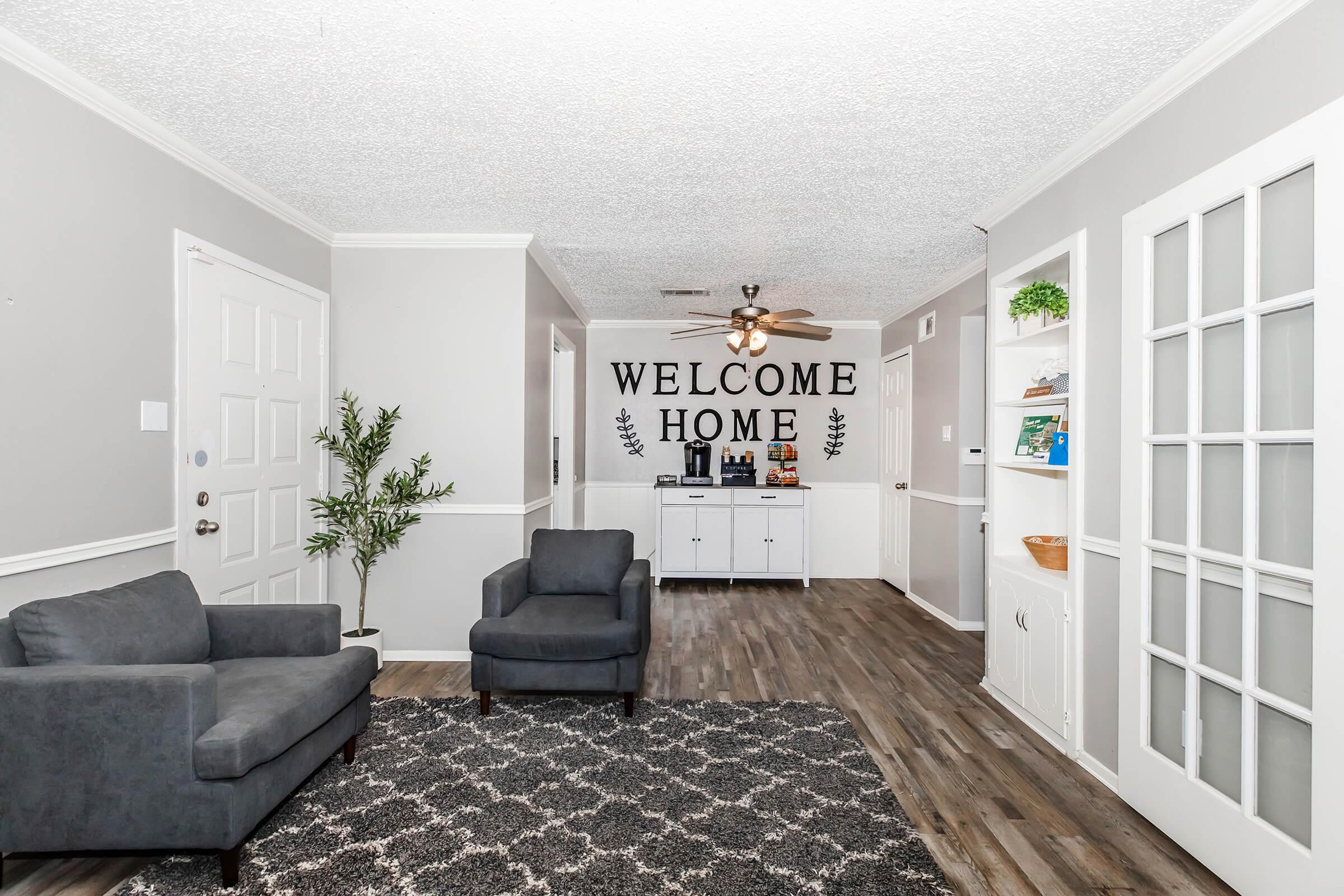 Cozy living space featuring grey walls, a welcome home wall decoration, a small seating area with a dark grey couch and armchair, and a wooden floor. A decorative plant sits in the corner, while shelves and a cabinet display books and decor on the adjacent wall.
